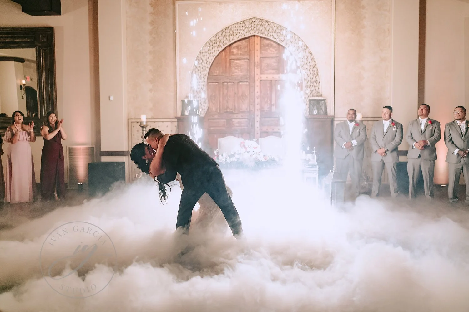 A couple dances in a cloud of fog during their wedding reception, surrounded by five groomsmen in suits and five bridesmaids, in a decorated ballroom.