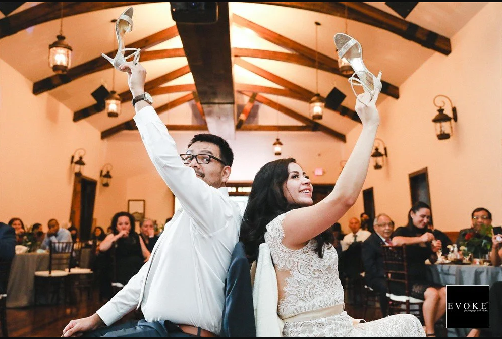 A couple is sitting back to back at a wedding or celebration, each holding up a high-heeled shoe. The man is wearing glasses and a white shirt, while the woman is dressed in a lace dress. They are smiling and engaging in a shoe game, with guests seated at tables watching in the background.