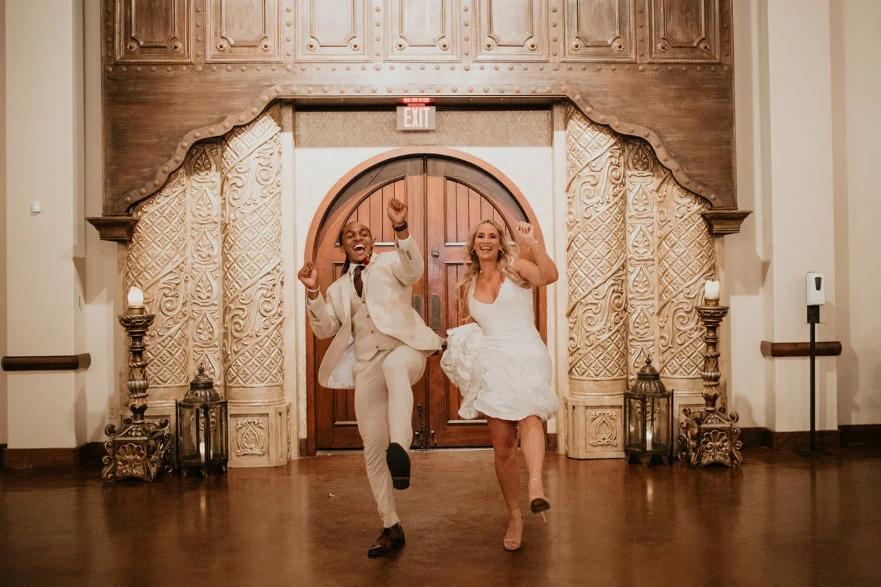 Two people in wedding attire dancing and celebrating in a decorated hall.