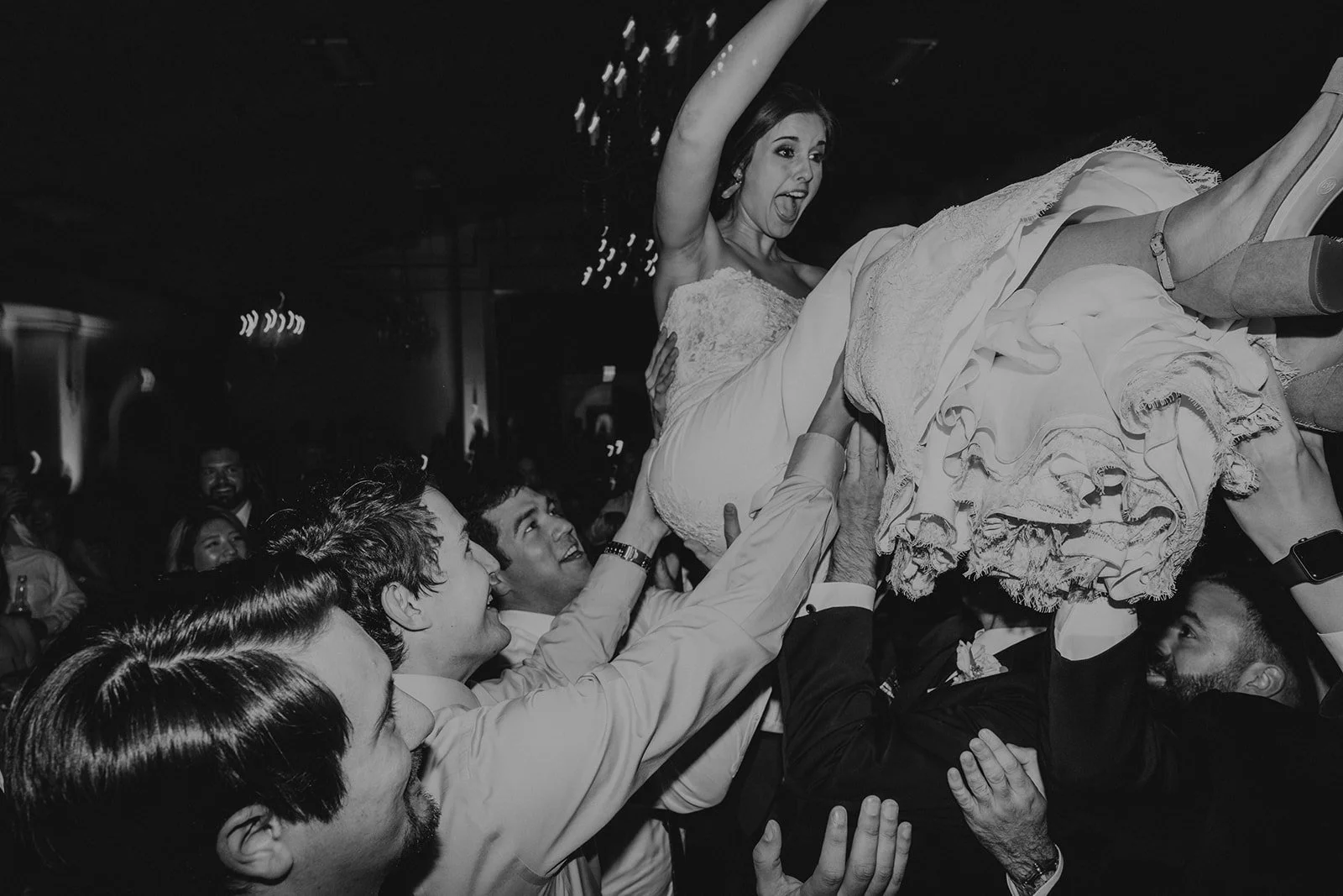 A bride being lifted by a group of people during her wedding celebration. The bride is smiling and excited, wearing a wedding dress. The scene is lively with friends and family in the background.