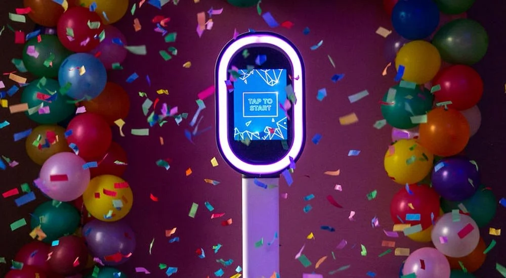 Colorful balloons and confetti surround a neon-lit interactive screen with the message 'Tap to Start' on a dark background.