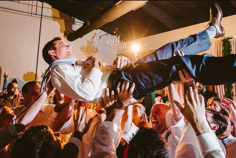 Man being lifted and crowd surfing at a celebration or wedding reception, smiling and laughing surrounded by people.