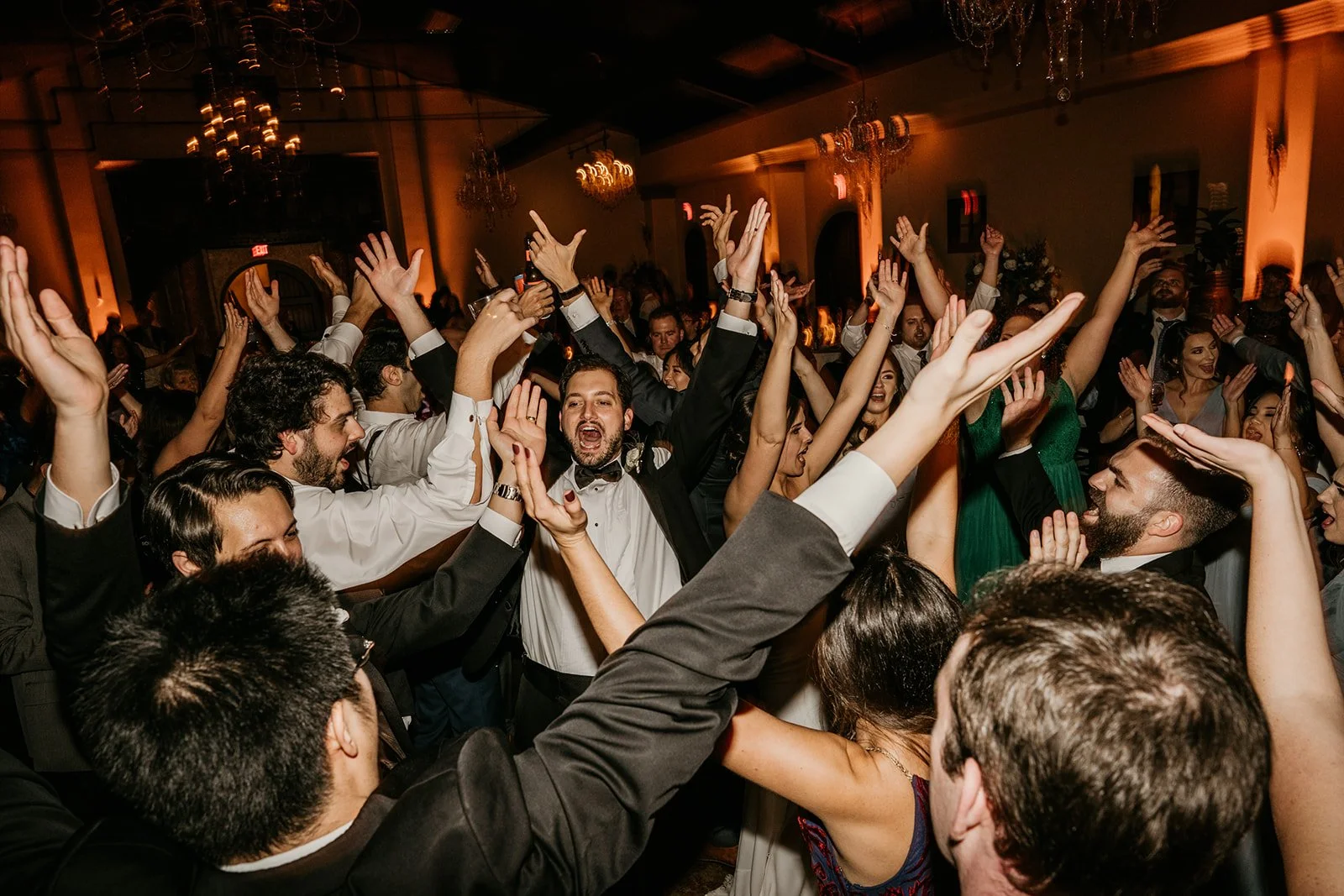 People dancing and celebrating at a lively, elegant indoor party with warm lighting and chandeliers.