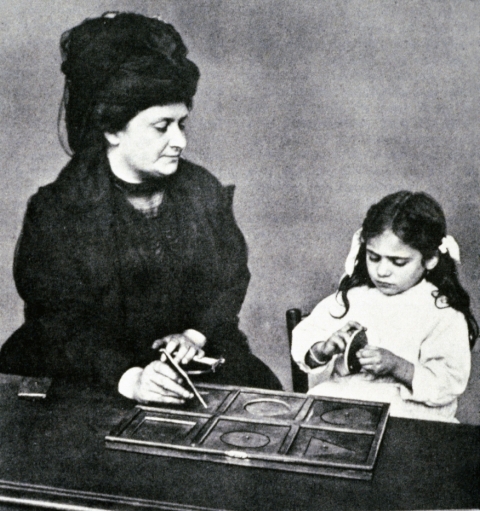 Dr. Maria Montessori modeling with a child how to use the metal insets, Early Children's House, Italy.