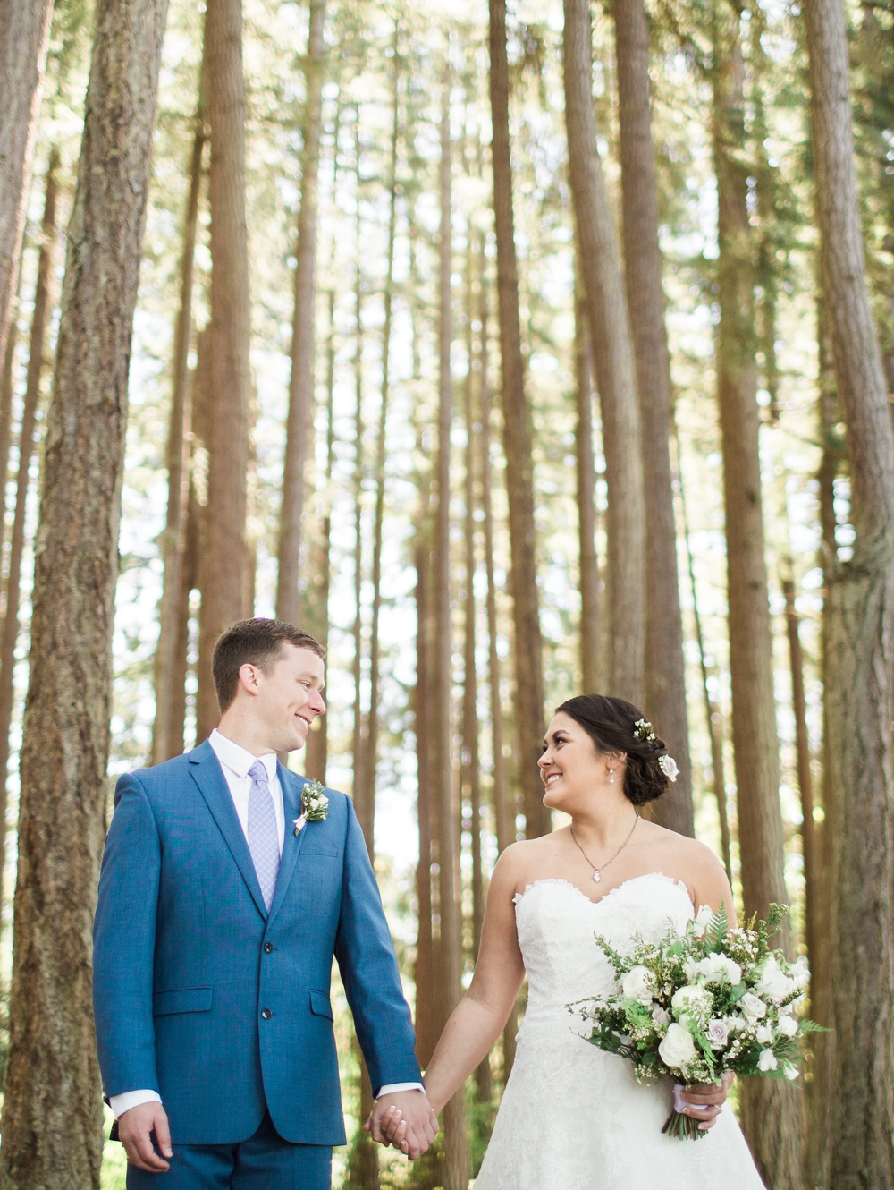  bride and groom holding hands and holding white bouquet in front of tall evergreen trees 