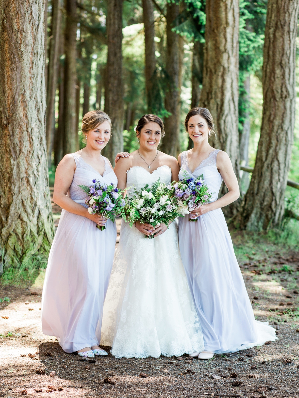  bride and brides maids in evergreen trees holding white and lavender spring bouquets 