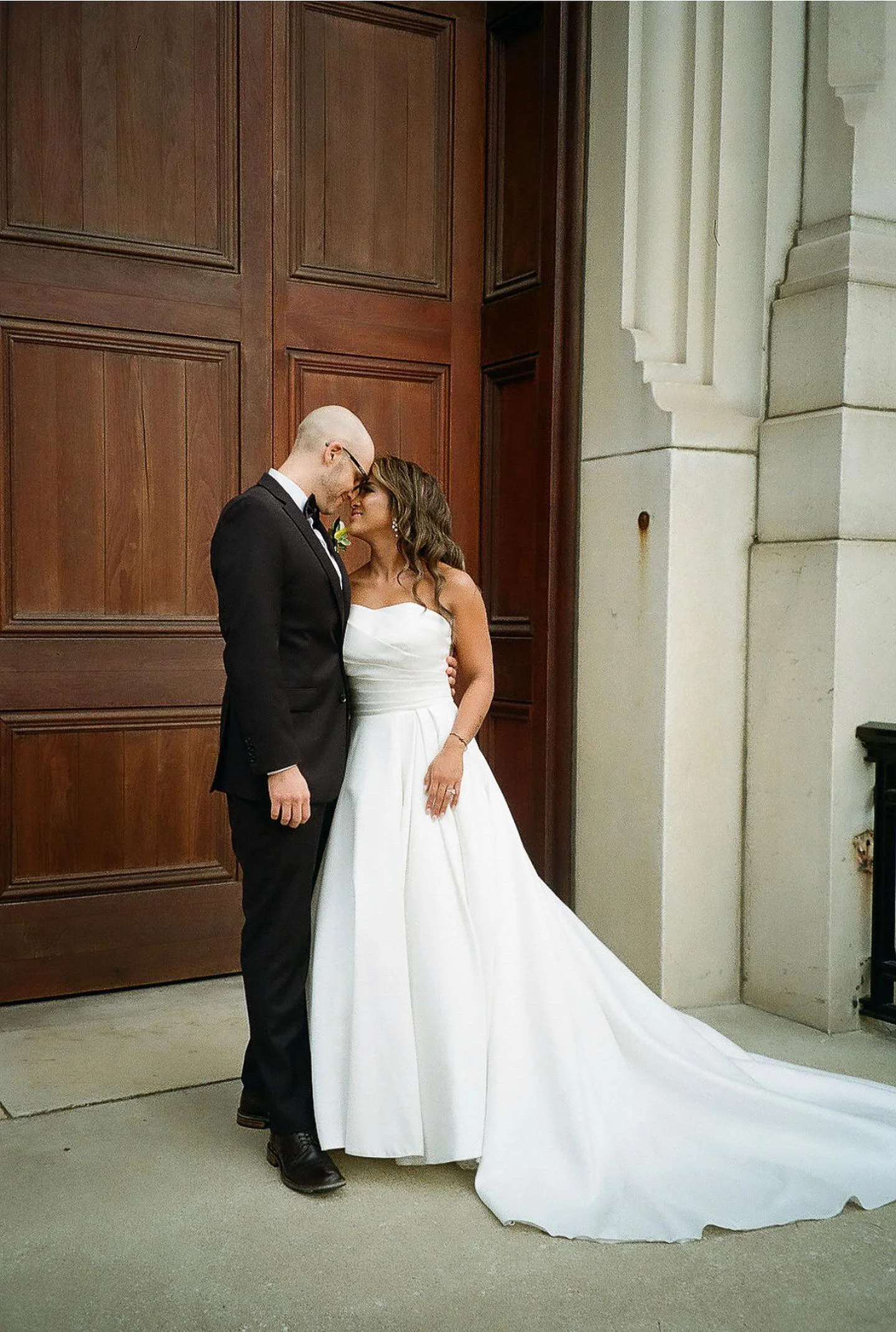A married couple stands closely in front of large wooden doors. The bride wears a long white gown, and the groom is in a black suit. They appear affectionate and are leaning towards each other.