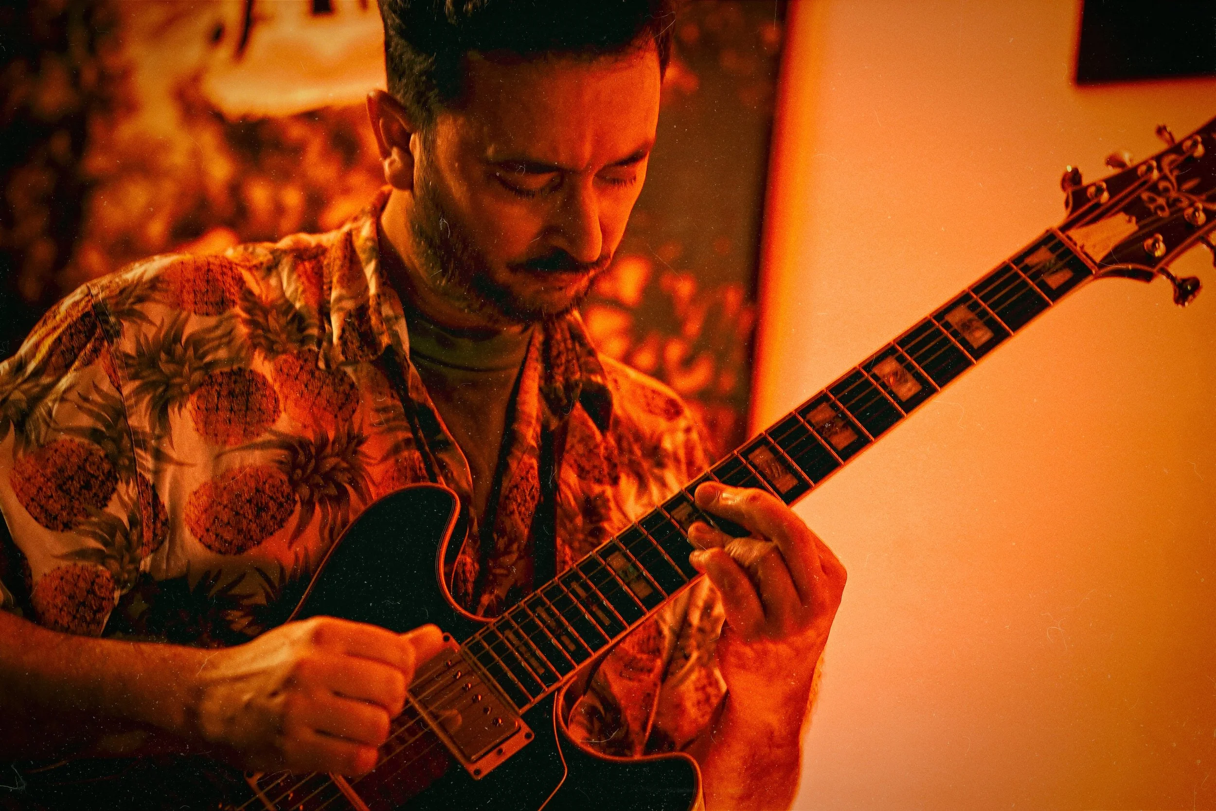 A man with dark hair and a beard playing an electric guitar. He is wearing a colorful pineapple patterned shirt and appears to be immersed in his music, with a warm, orange-lit background.