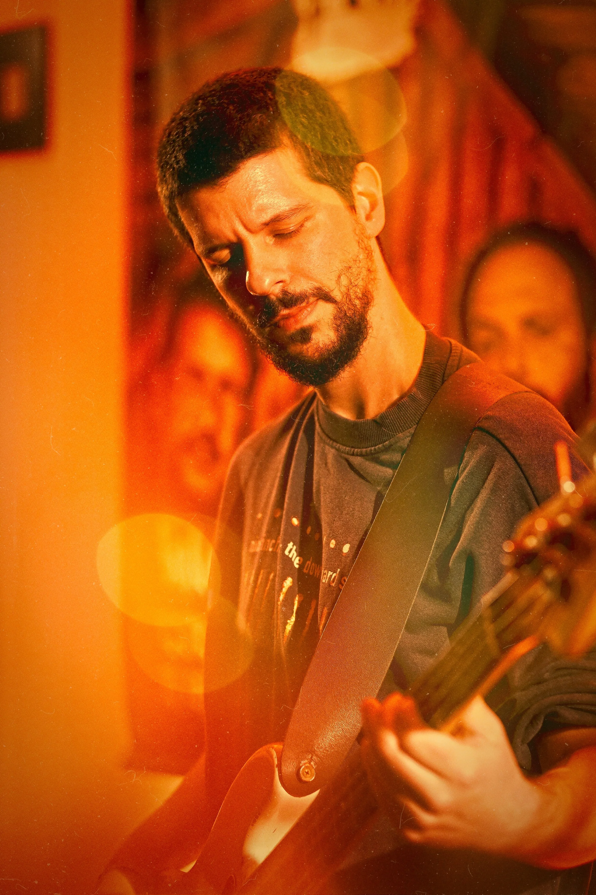 A man with dark hair and beard playing an electric guitar in a dimly lit room with warm yellow and orange lighting, with artwork on the walls behind him.