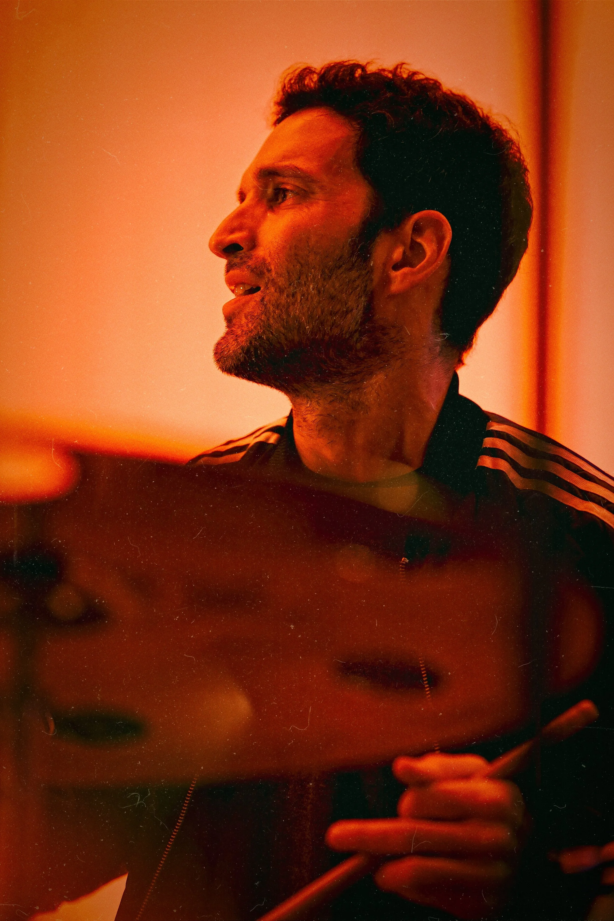 Side profile of a man with short curly hair and a beard, wearing a black jacket with white stripes, holding a pen or stylus, illuminated by warm orange lighting.