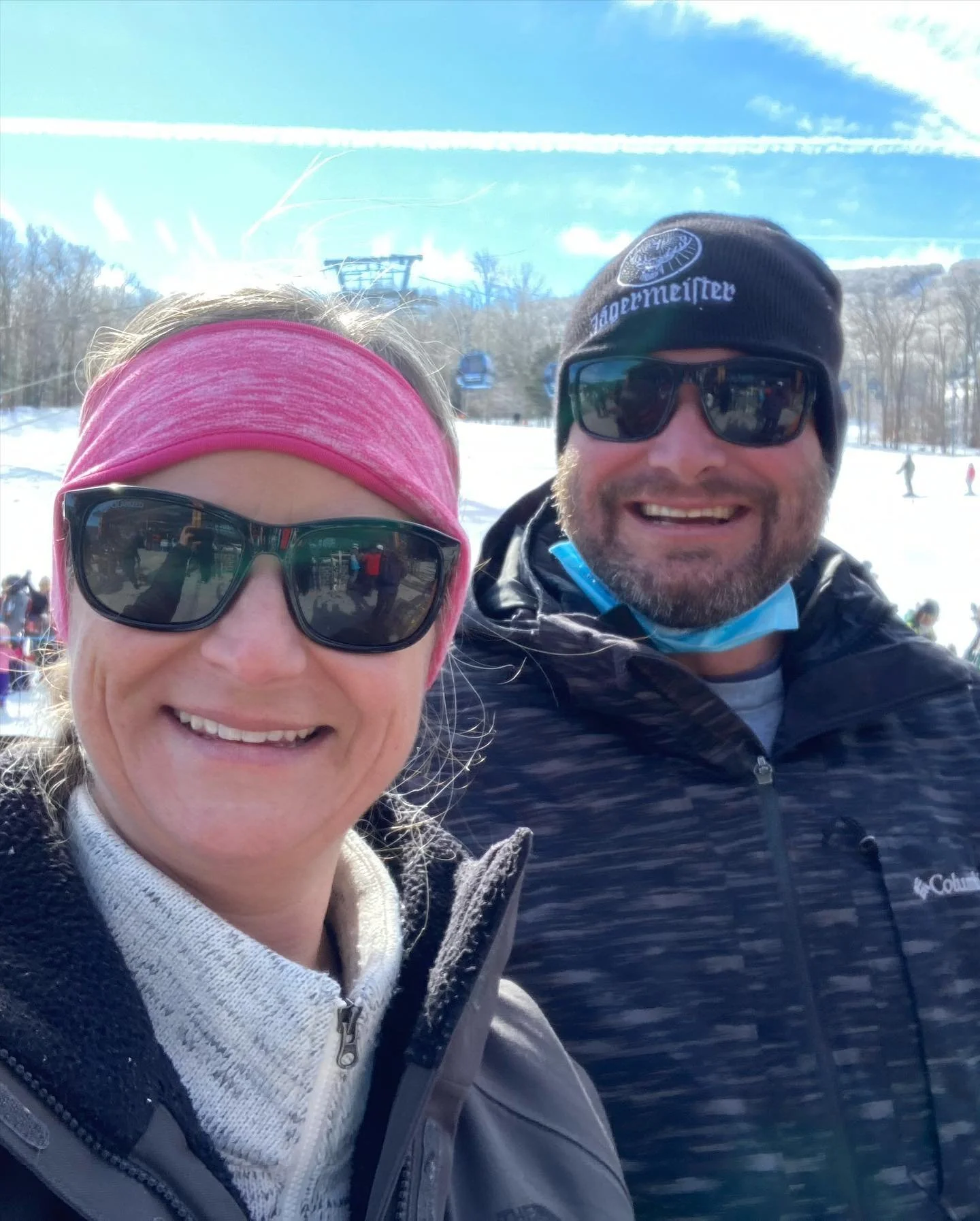 Smiling man and woman taking a selfie at a snowy ski resort, both wearing sunglasses and winter jackets. The woman has a pink headband, and the man has a black beanie and sunglasses with a jacket. Ski lifts and other skiers are visible in the background.