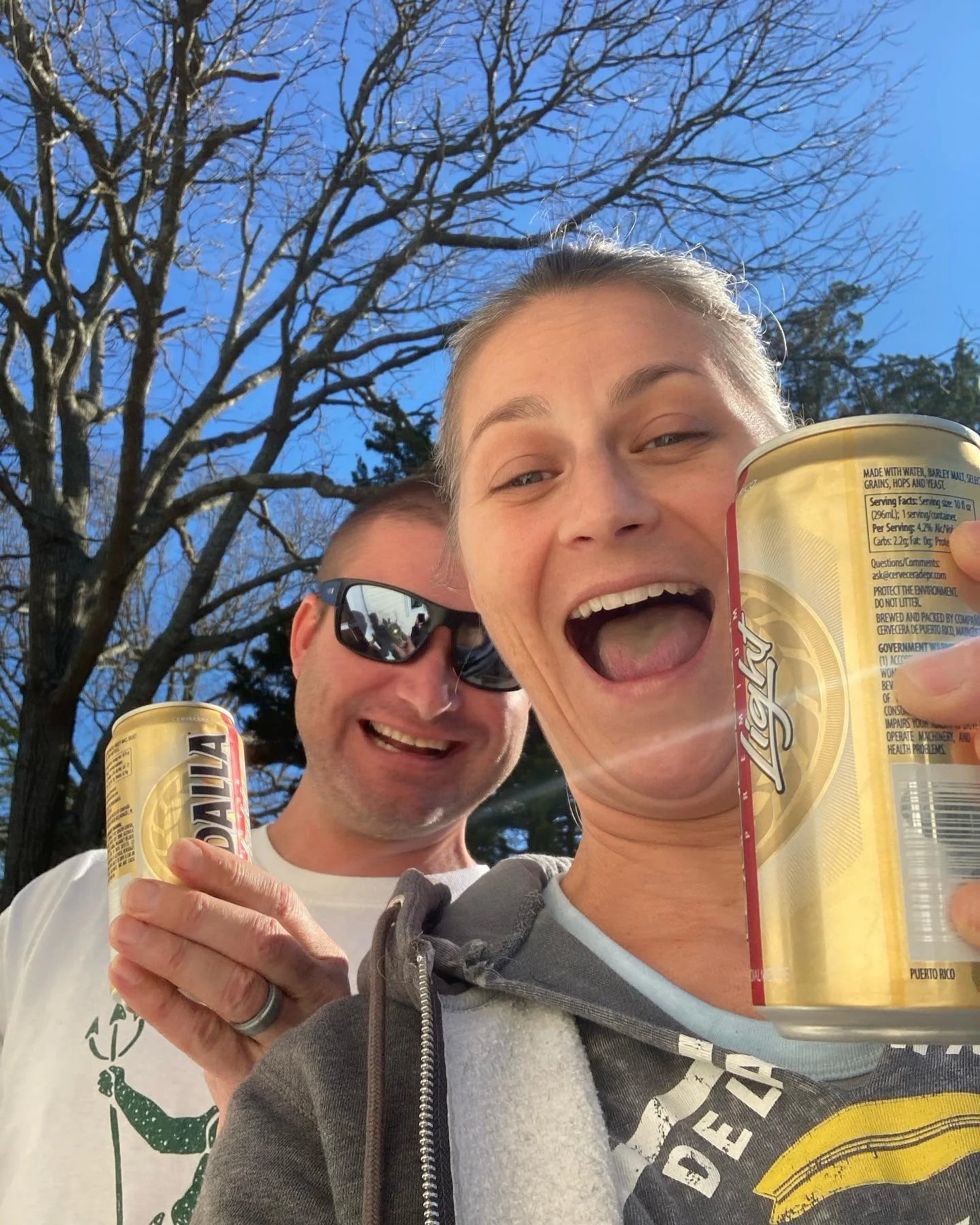 A woman and a man outdoors on a sunny day, smiling and holding canned beverages. The woman is in the foreground, taking a selfie, holding a yellow can, and the man is behind her wearing sunglasses, also holding a canned beverage.