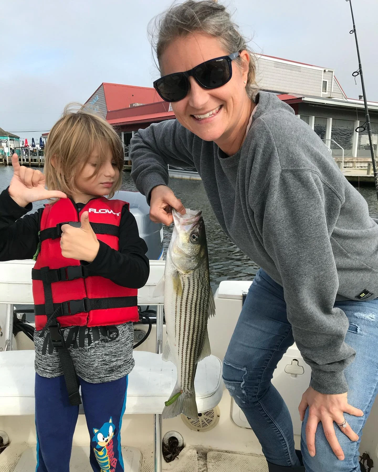 A woman and a child on a boat holding a large striped bass fish. The woman is smiling and wearing sunglasses, the child is giving a thumbs-up and wearing a red life jacket. There are waterfront buildings in the background.