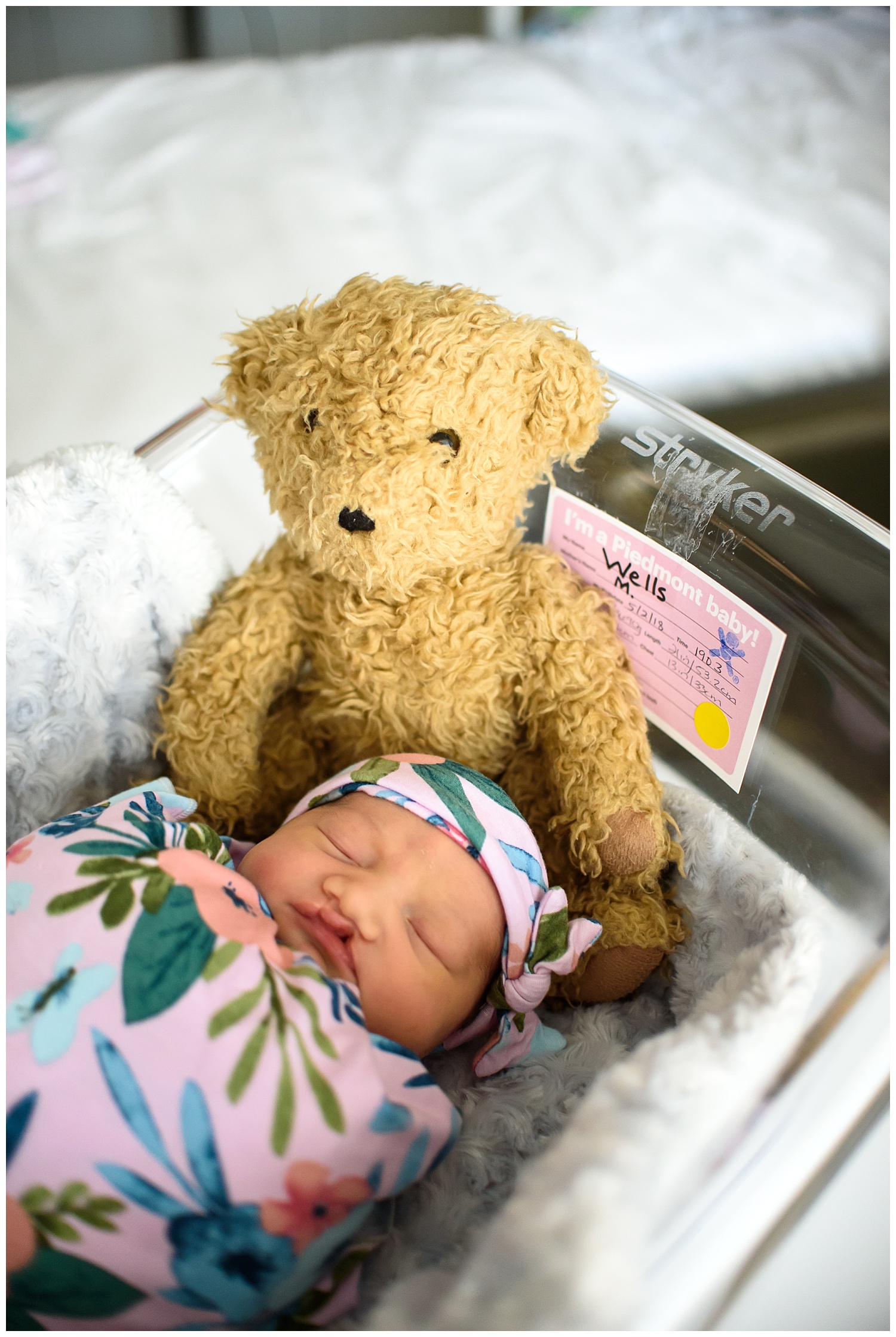 this is an image of a newborn baby girl laying in a hospital bassinet. she is sleeping and there is a family teddy bear next to her in the bassinet.
