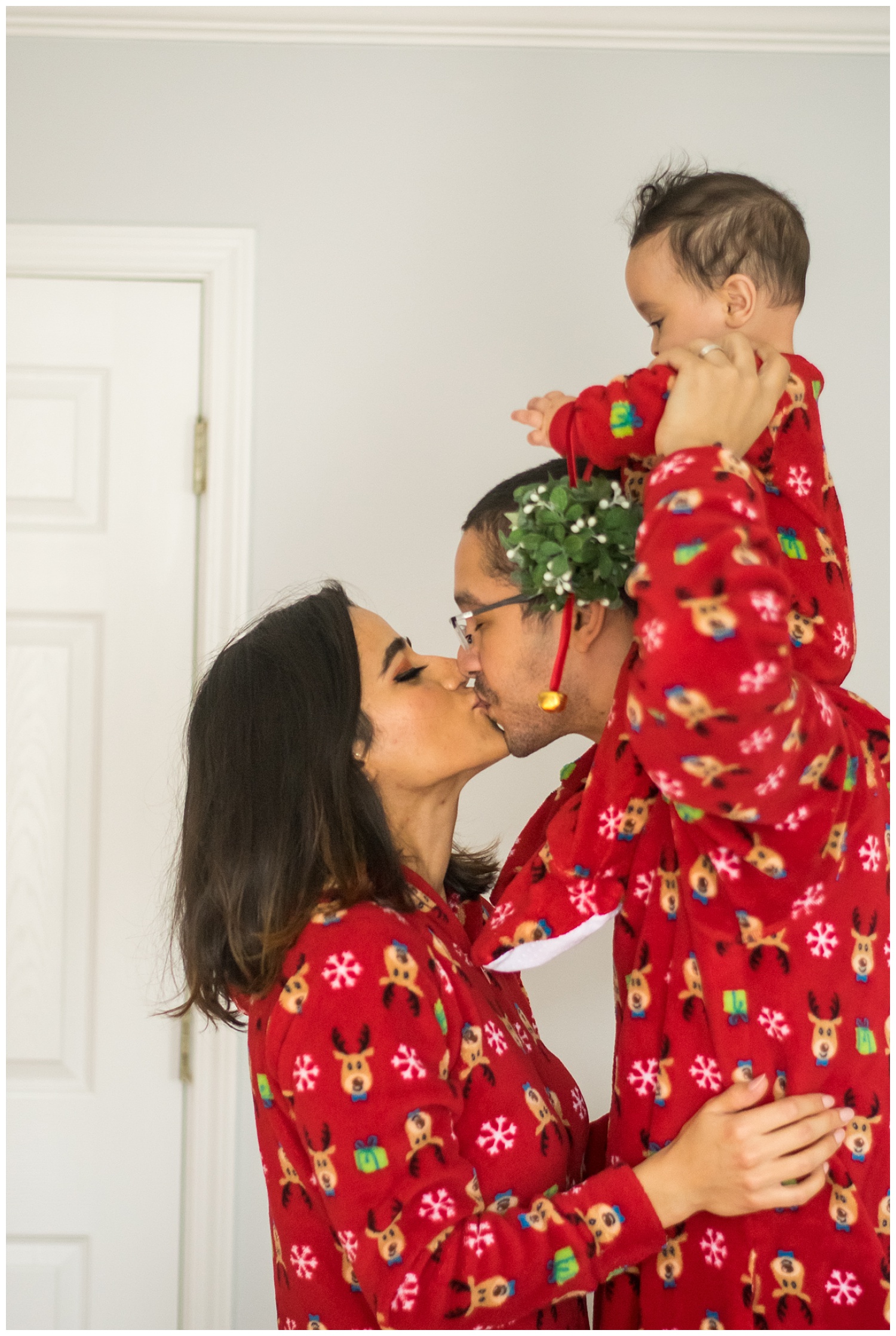 this is an image of a family in matching christmas pajamas. dad is holding the six month boy on his shoulders and mom and dad are under the mistletoe that the baby is holding