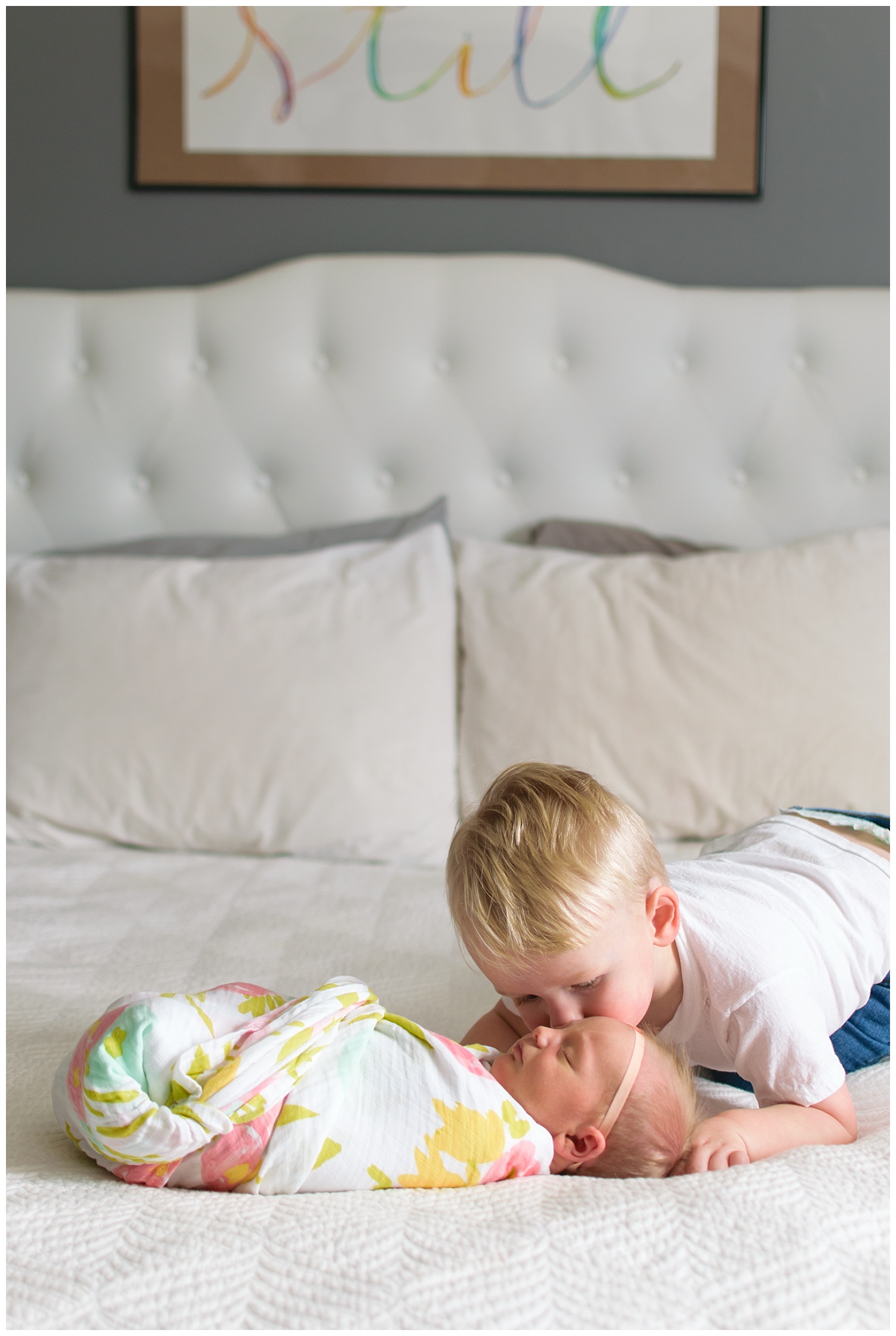 this image was taken during a lifestyle newborn family session. the baby girl is laying on the bed and her brother is kissing her cheek.