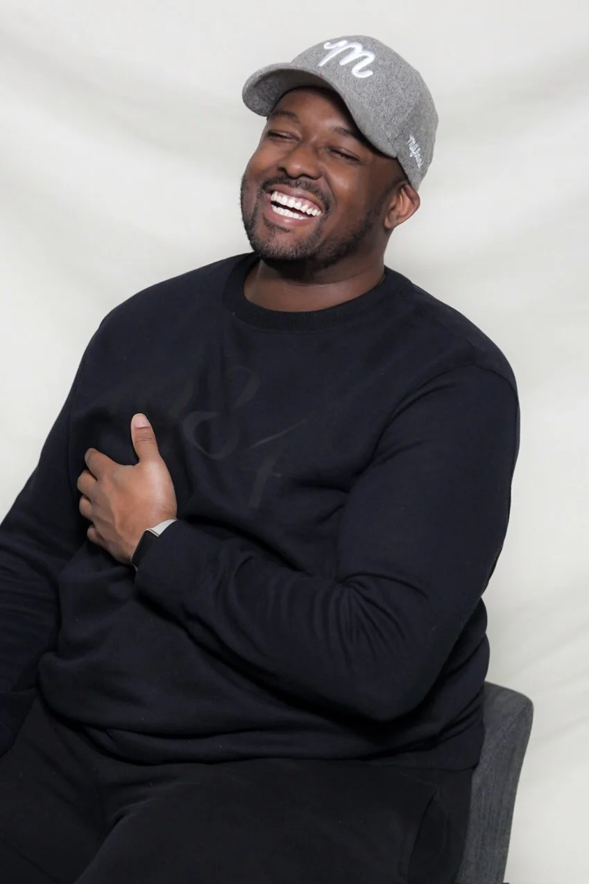 Man wearing a gray cap and black shirt, smiling and touching his chest, sitting against a plain light background.