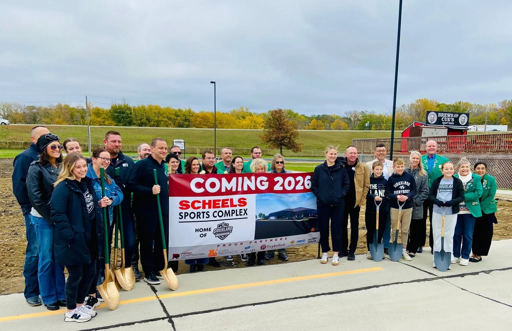 Group of people outdoors at a groundbreaking ceremony for a new sports complex, holding shovels, with a banner that reads "Coming 2026 SCL Sports Complex."