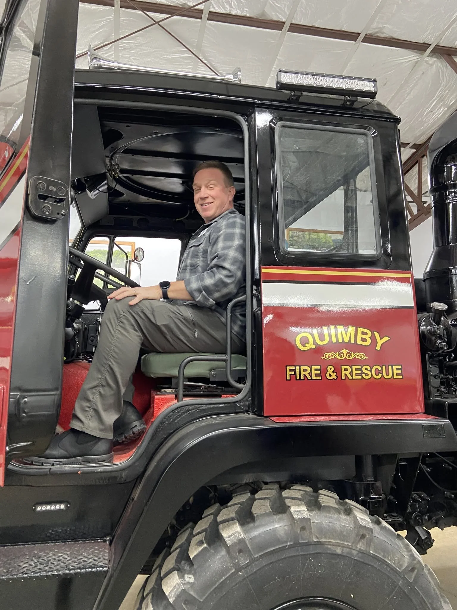 A smiling man sitting inside a fire and rescue vehicle, labeled 'QUIMBY FIRE & RESCUE,' in an indoor setting.