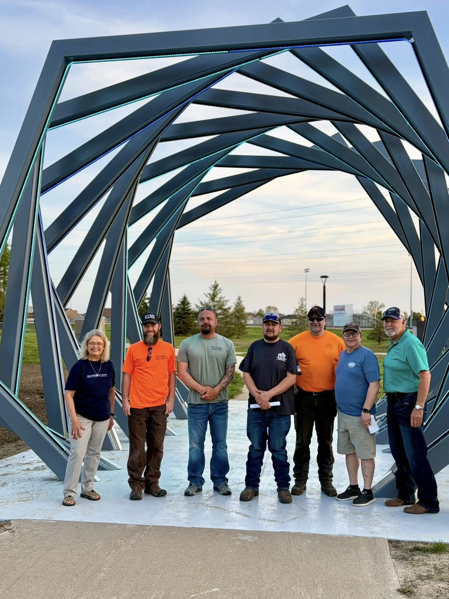Group of seven people standing outdoors under a metal arch structure, smiling at the camera, with trees and a field in the background.