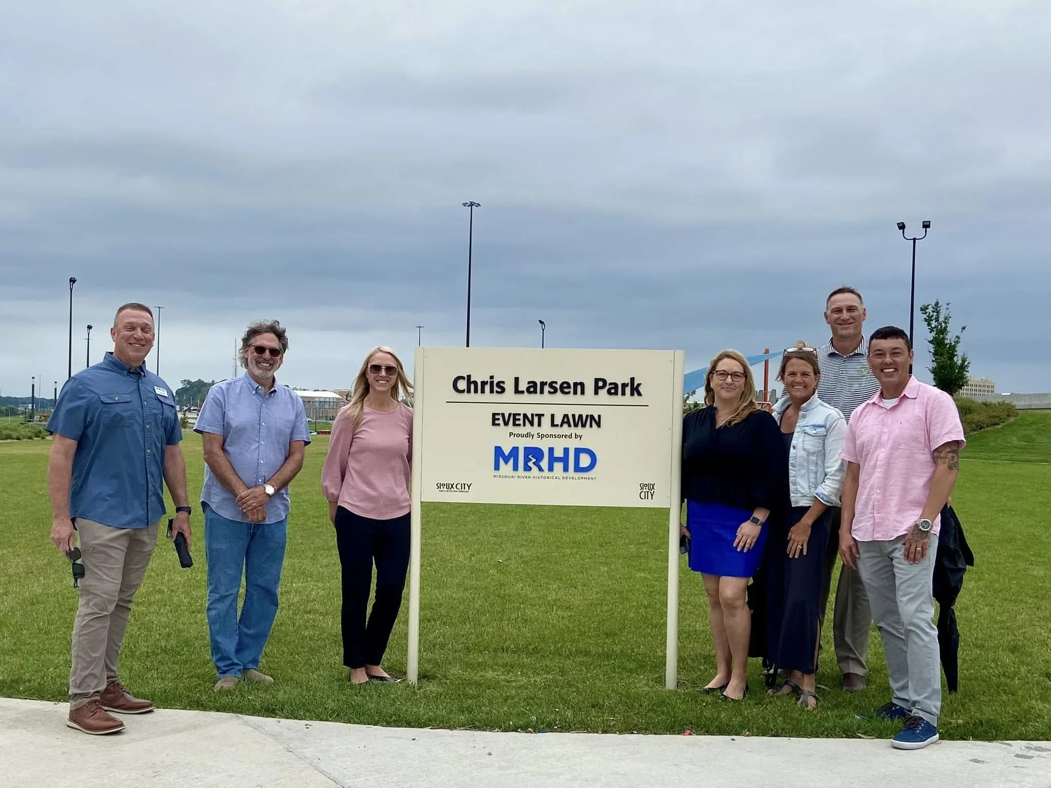 Group of people standing outdoors at Chris Larsen Park event lawn, smiling and posing for the photo, with cloudy sky and green grass in the background.