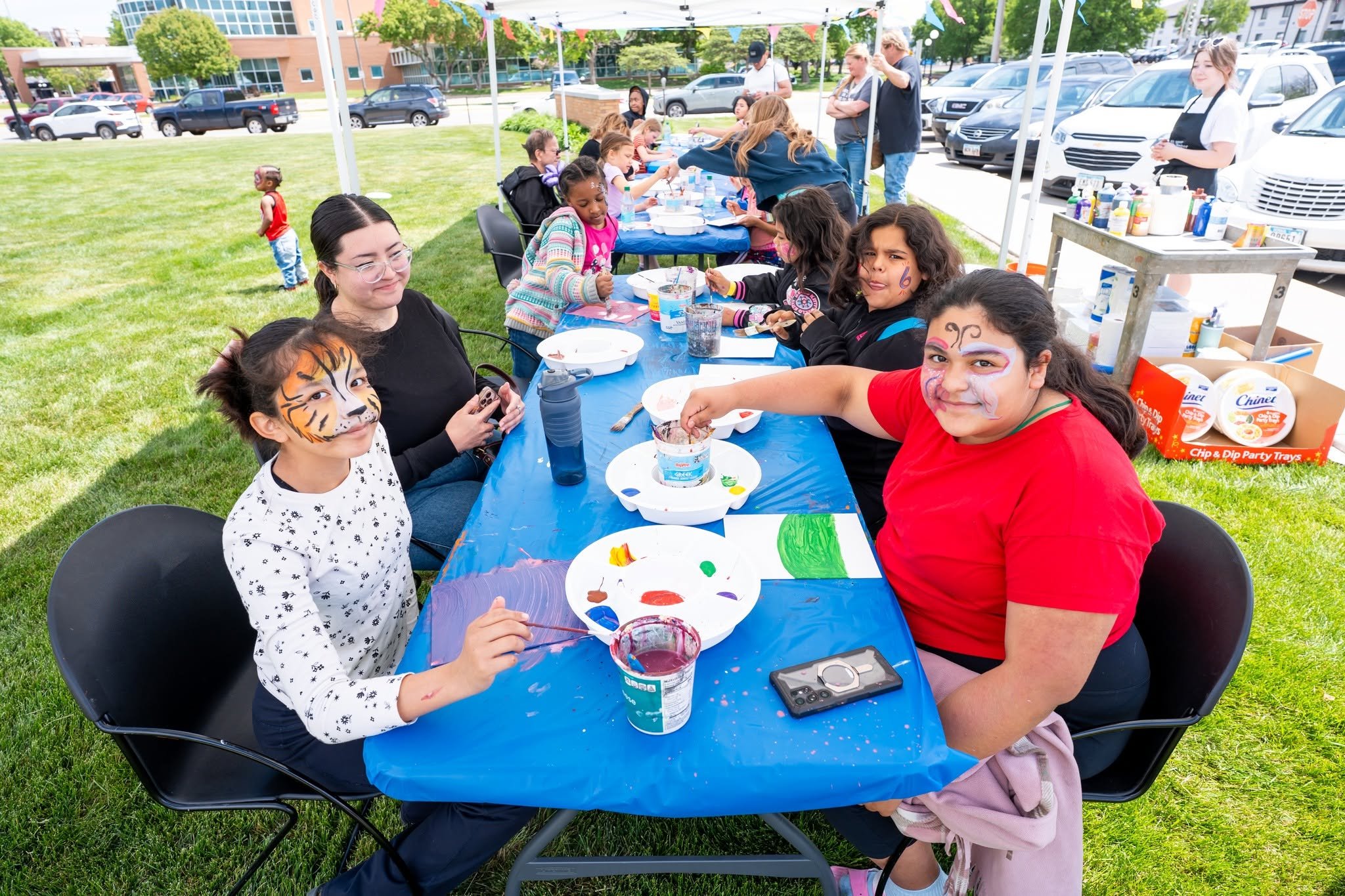 Children and adults at an outdoor painting birthday party under a tent on a grassy area, with parking lot in the background.