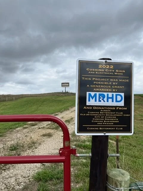 A signboard in Cushing City noting 2022 city improvements and electrical work, supported by grants and donations, with a red barrier in the foreground and a grassy hill under cloudy skies.