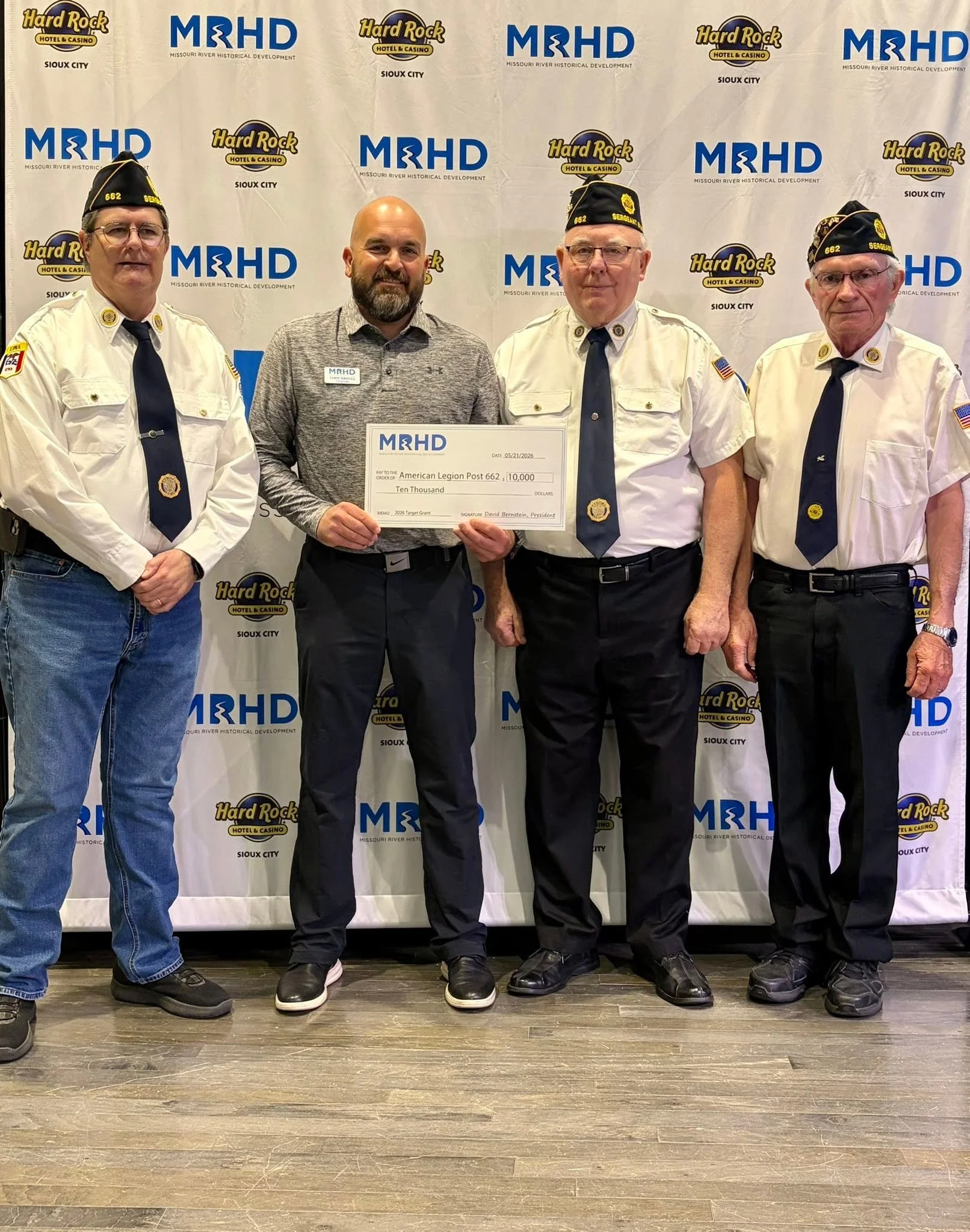 Four men standing in front of a backdrop with logos of Hard Rock Hotel & Casino Sioux City and Missouri River Historical Development. One man holds a large check for $10,000 made out to American Legion Post 662. Three men wear white shirts and black hats with gold insignia, the man with the check wears a gray shirt. They are all smiling or looking serious, standing on a wooden floor.
