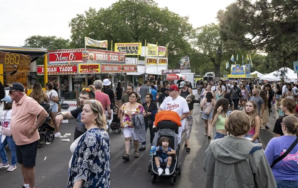 Crowd of people walking around at a fair or festival with food stalls and game booths in the background, surrounded by trees.