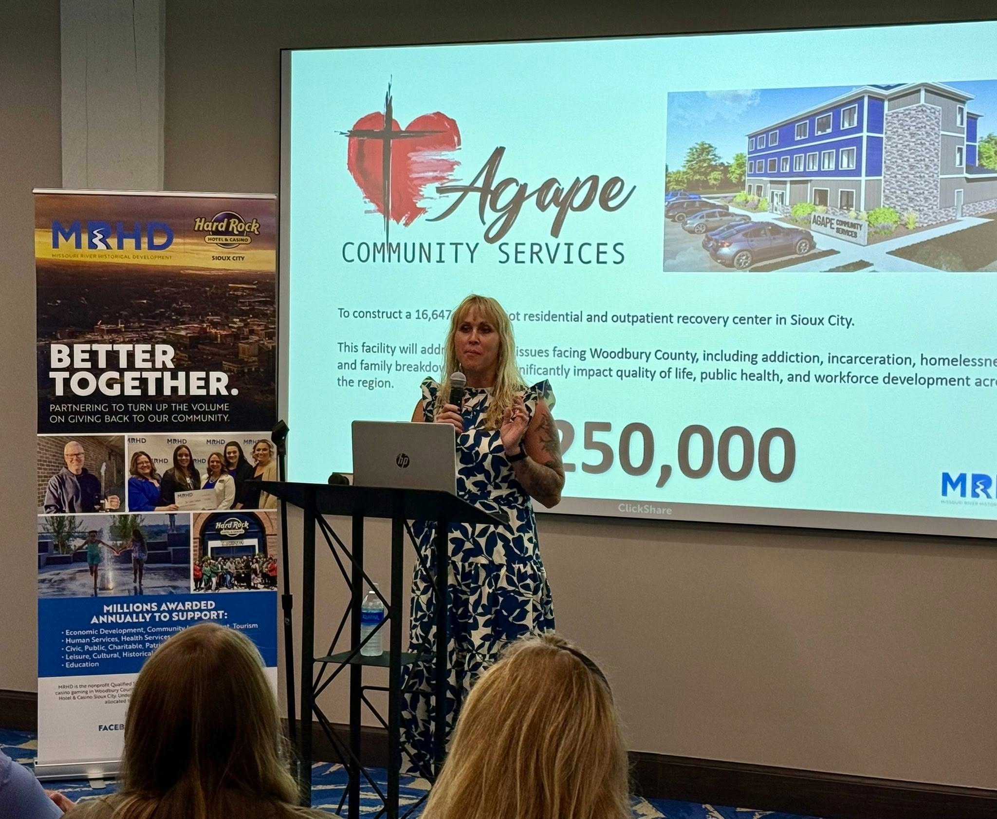 A woman speaking at a presentation about Agape Community Services, with a slide showing a building and parking lot, and banners related to Missouri River Historical Development and Hard Rock Hotel & Casino Sioux City in the background.
