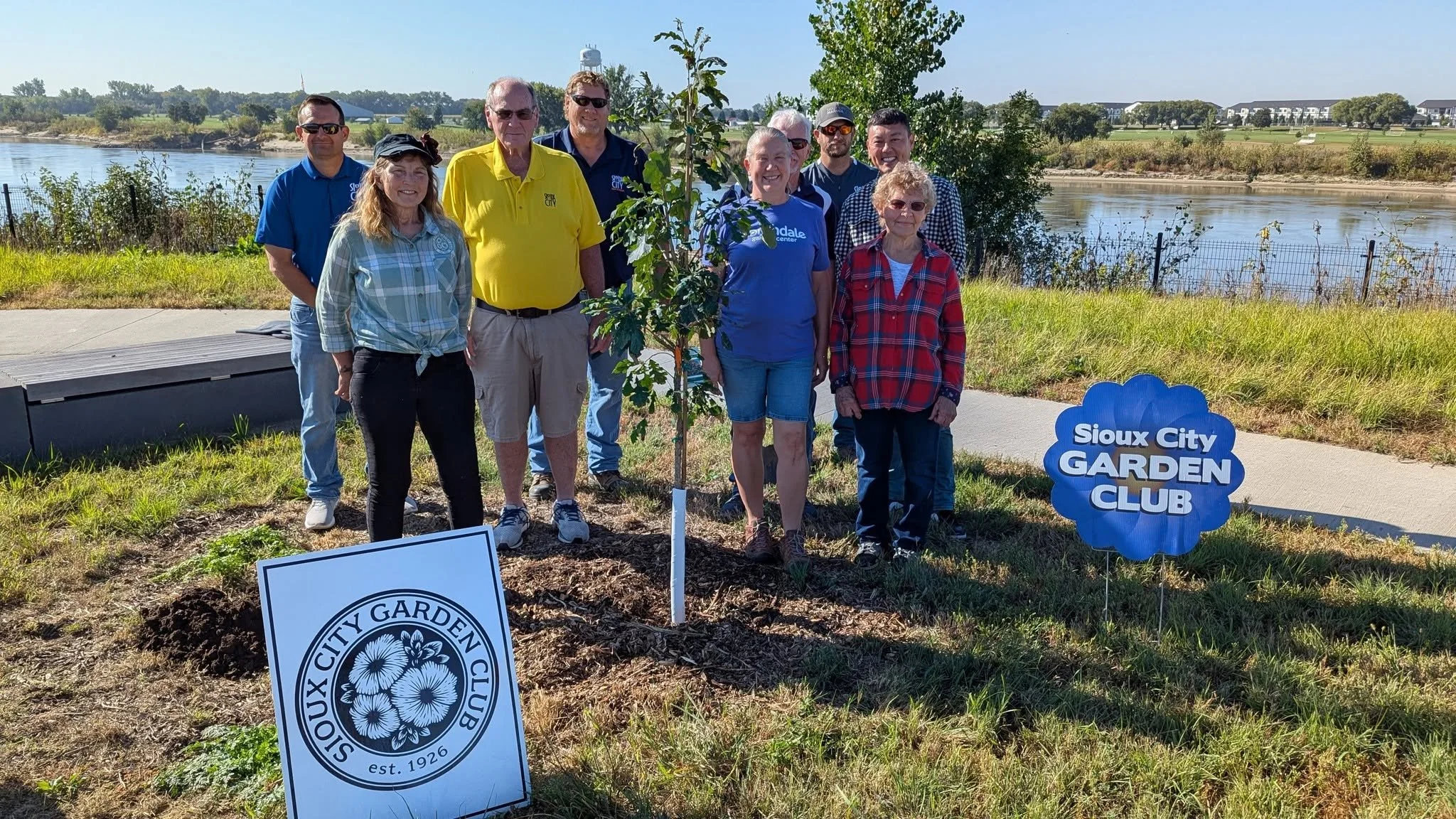 A group of people standing in front of a small tree at a Sioux City Garden Club event by a body of water. There are signs for the Sioux City Garden Club and Garden Center.