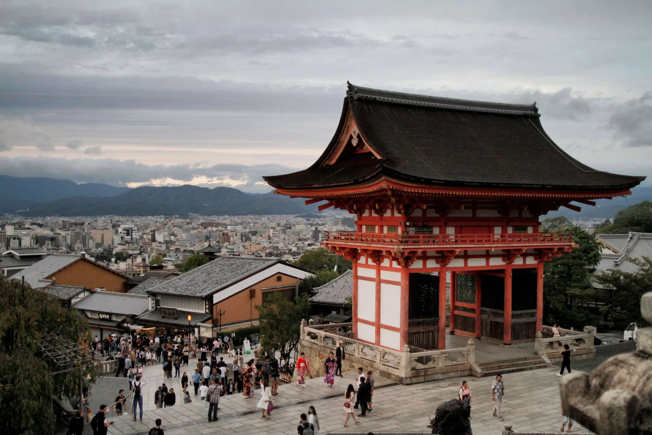 Traditional Japanese temple with a large, curved roof, red support beams, and multiple black tiles, overlooking a cityscape with a crowd of people walking on stone steps and paths in Kyoto