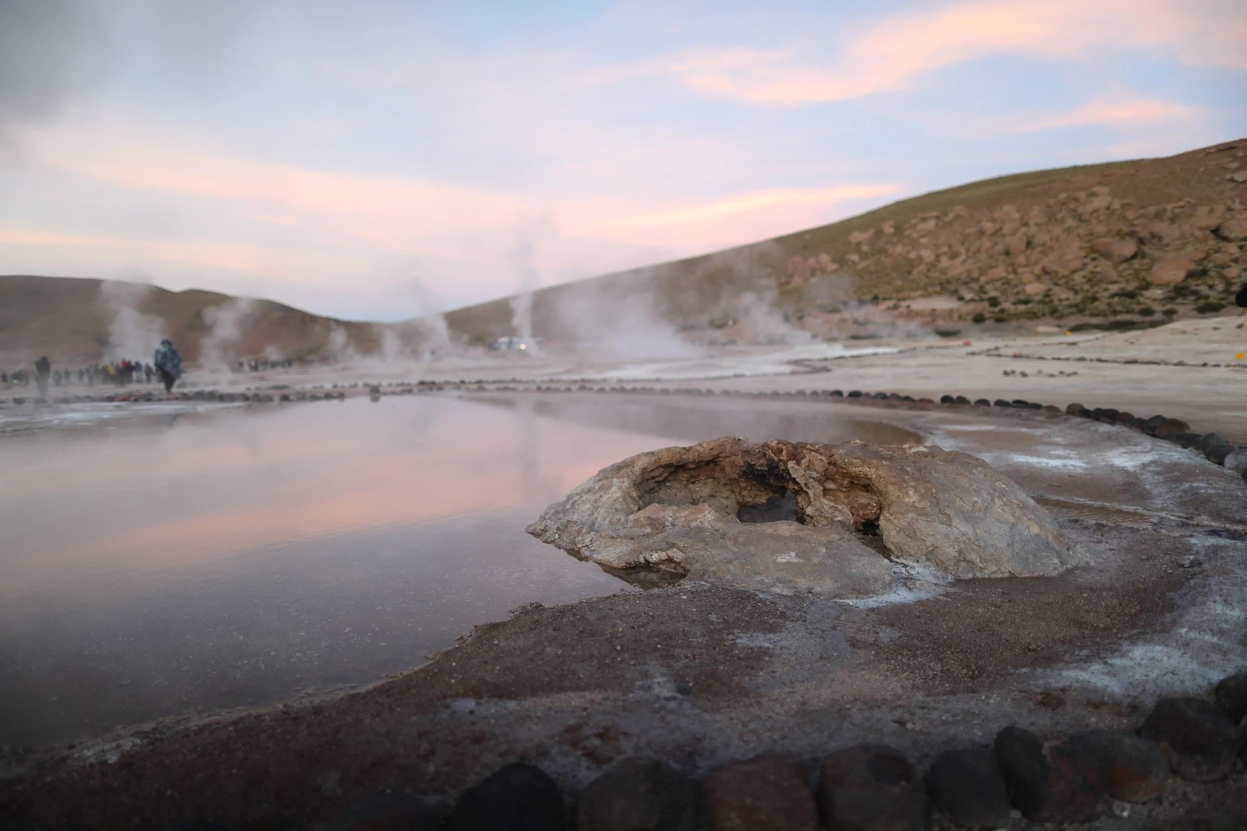 Geothermal hot springs with steam rising, rocky and barren terrain, hills in the background, and a group of people in the distance at sunrise, Chile's El Tatio geysers in Atacama