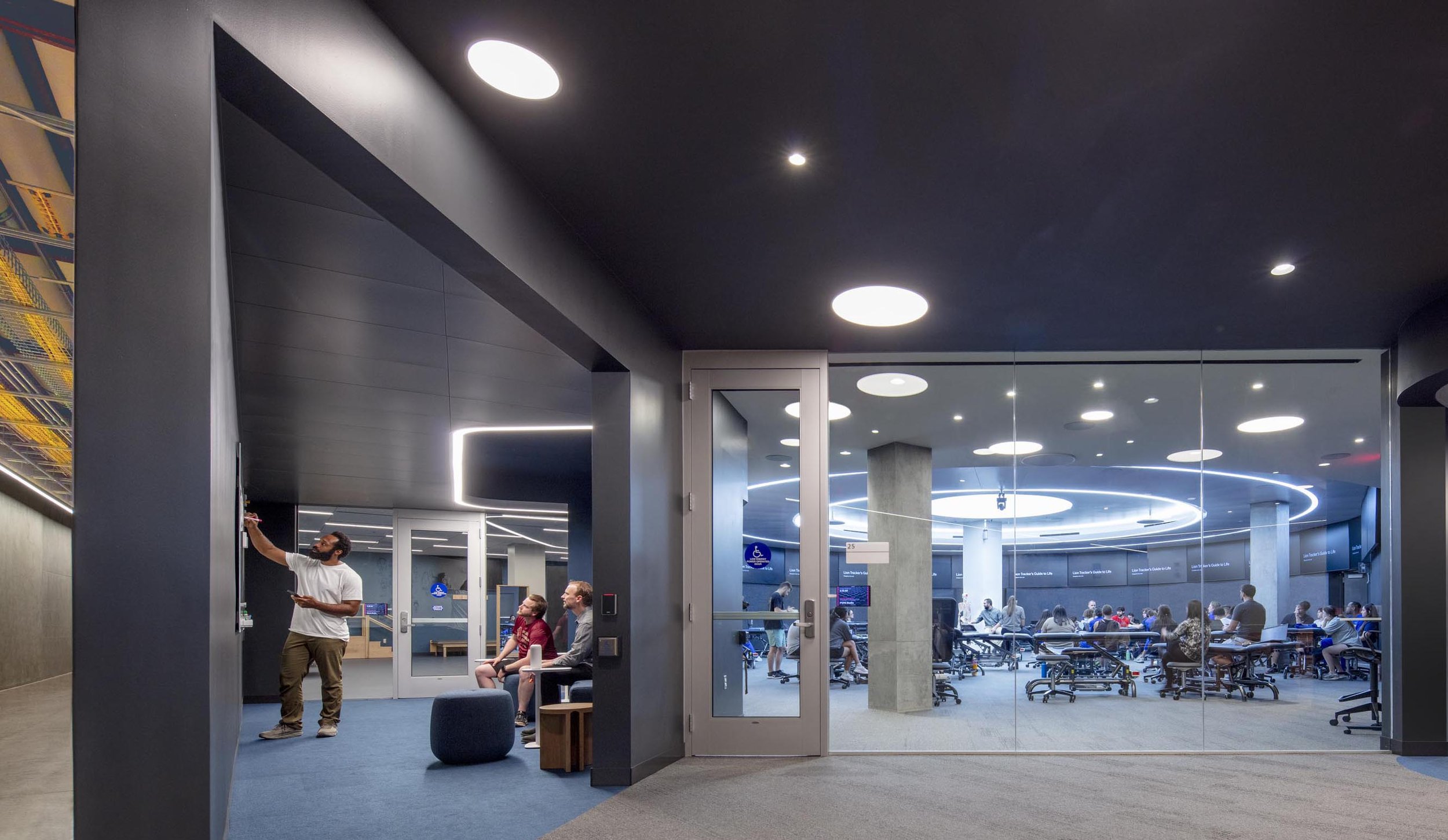 Student working in a classroom suite with modern lighting and seating areas, some sitting and some standing.