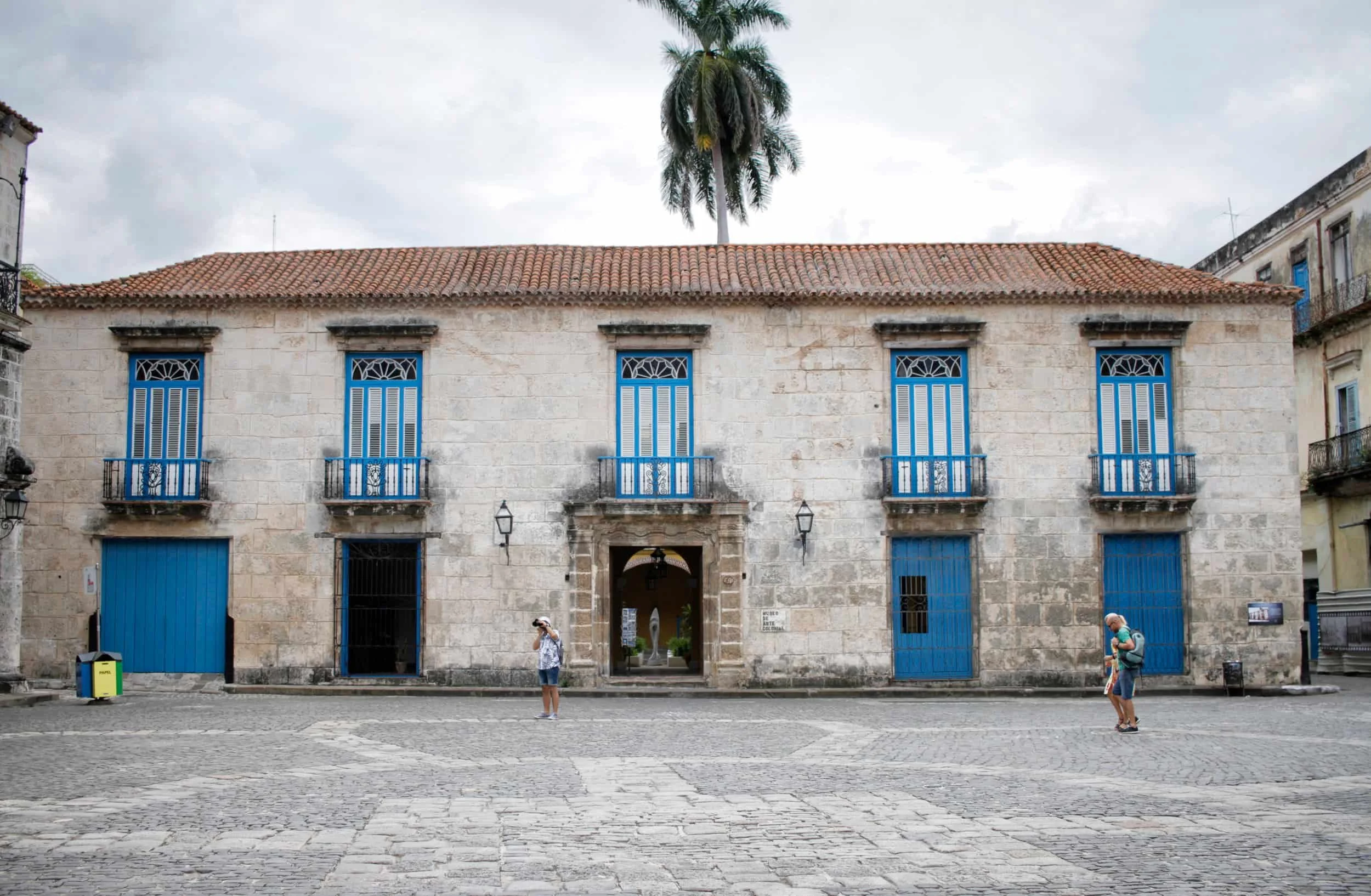 A historic stone building with blue doors and windows, a terracotta tile roof, a palm tree on top, and people walking in front in a Spanish colonial town square. Havana, Cuba.
