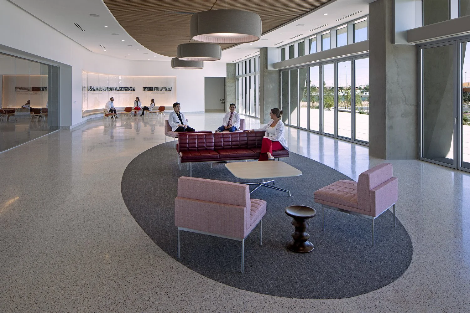 Modern university building waiting area with pink and red chairs, a black circular rug, large windows, and students in white coats seated and conversing.