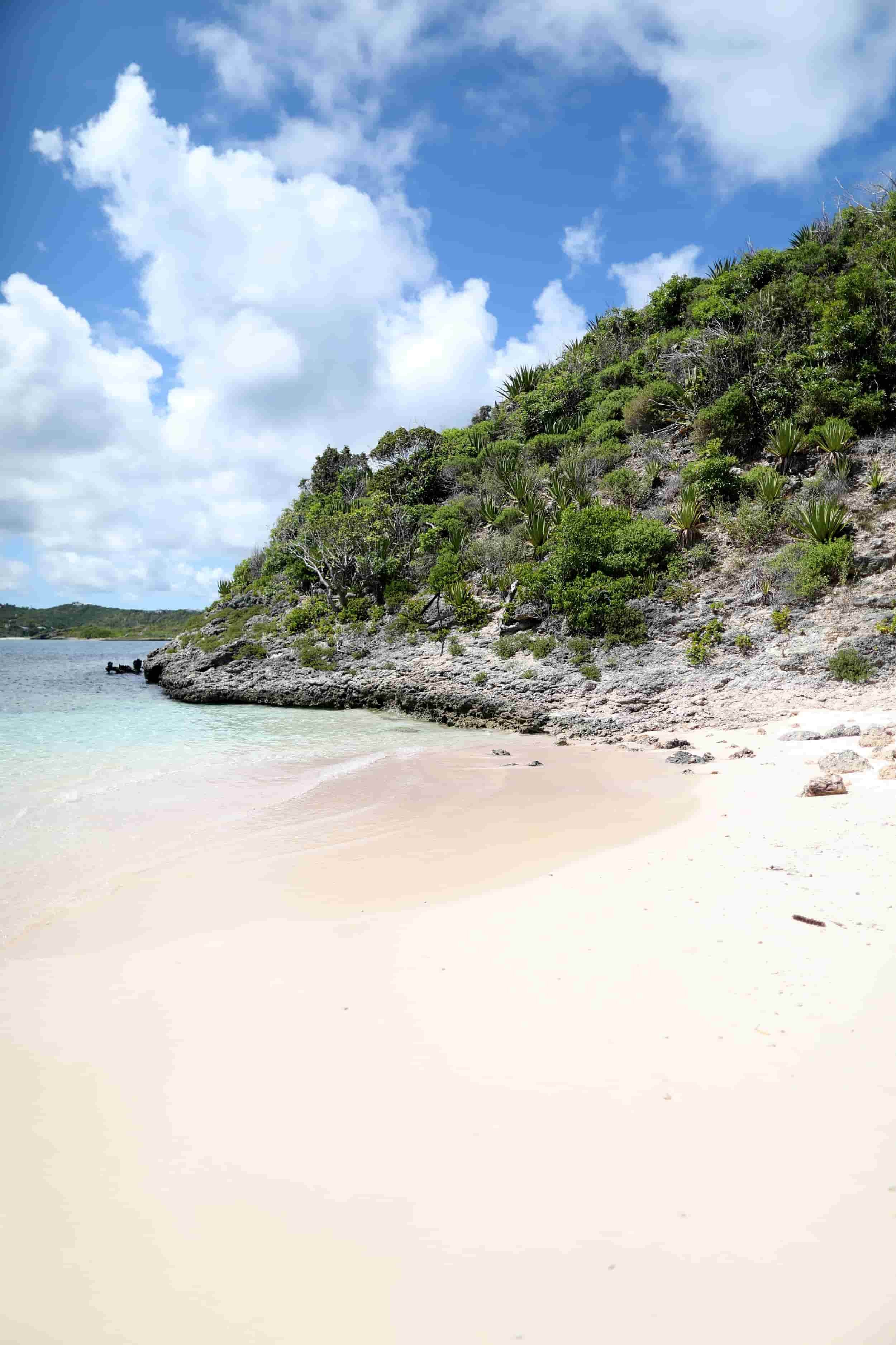 A tropical beach with white sand, calm turquoise water, lush green vegetation on a rocky hillside, and a partly cloudy blue sky.