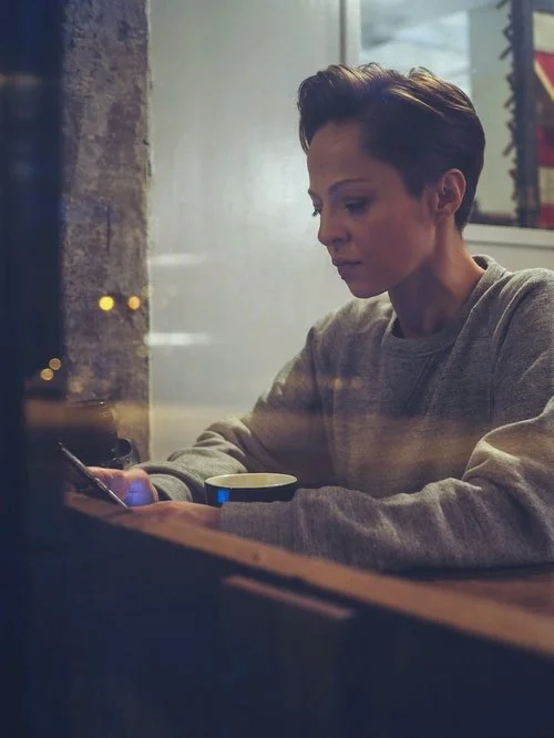 A woman with short hair sitting at a table in a cafe or restaurant, looking at her phone, with a coffee cup in front of her, seen through a window with reflections.