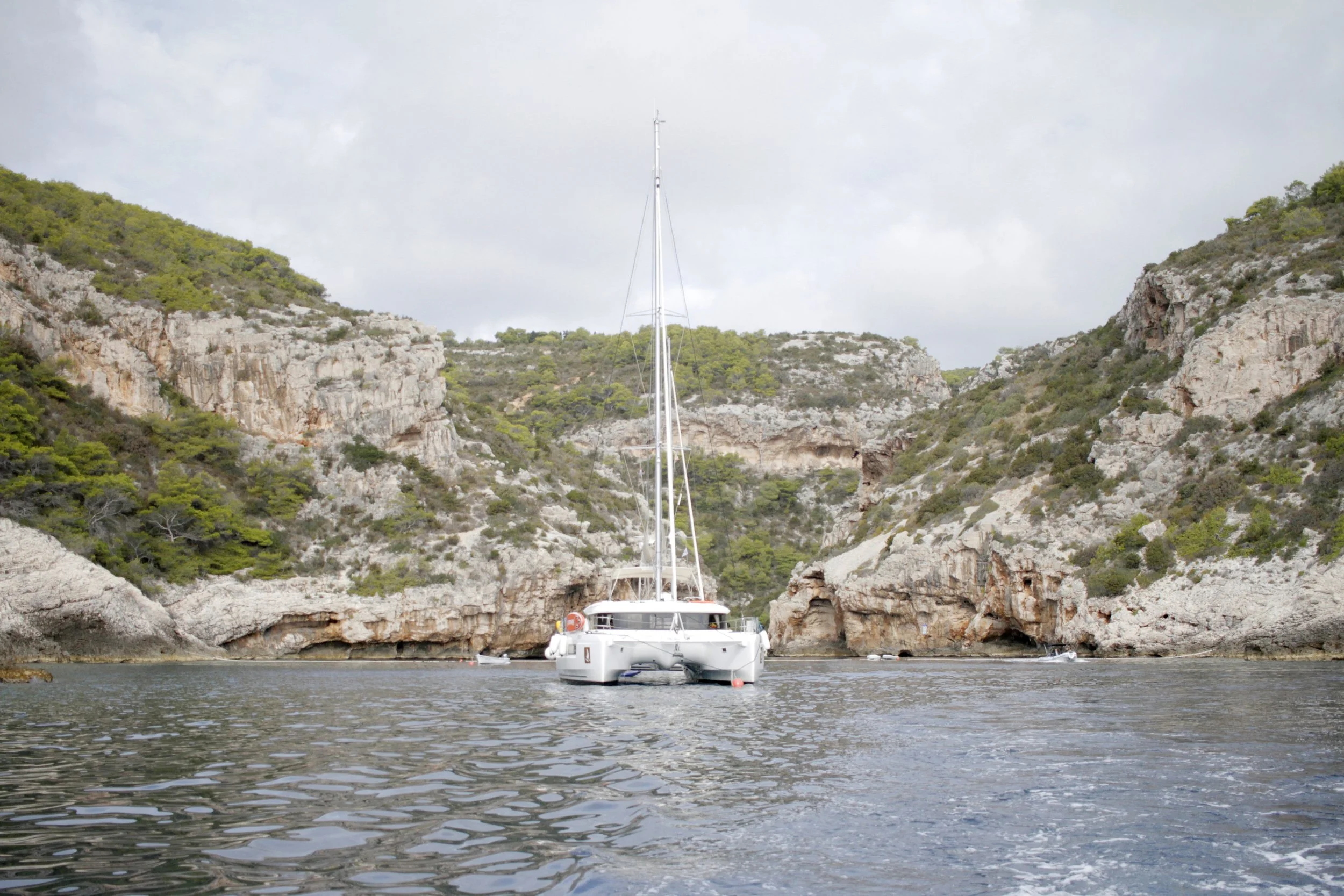 A white sailboat with a tall mast cruising through calm water near rocky cliffs with sparse green vegetation.