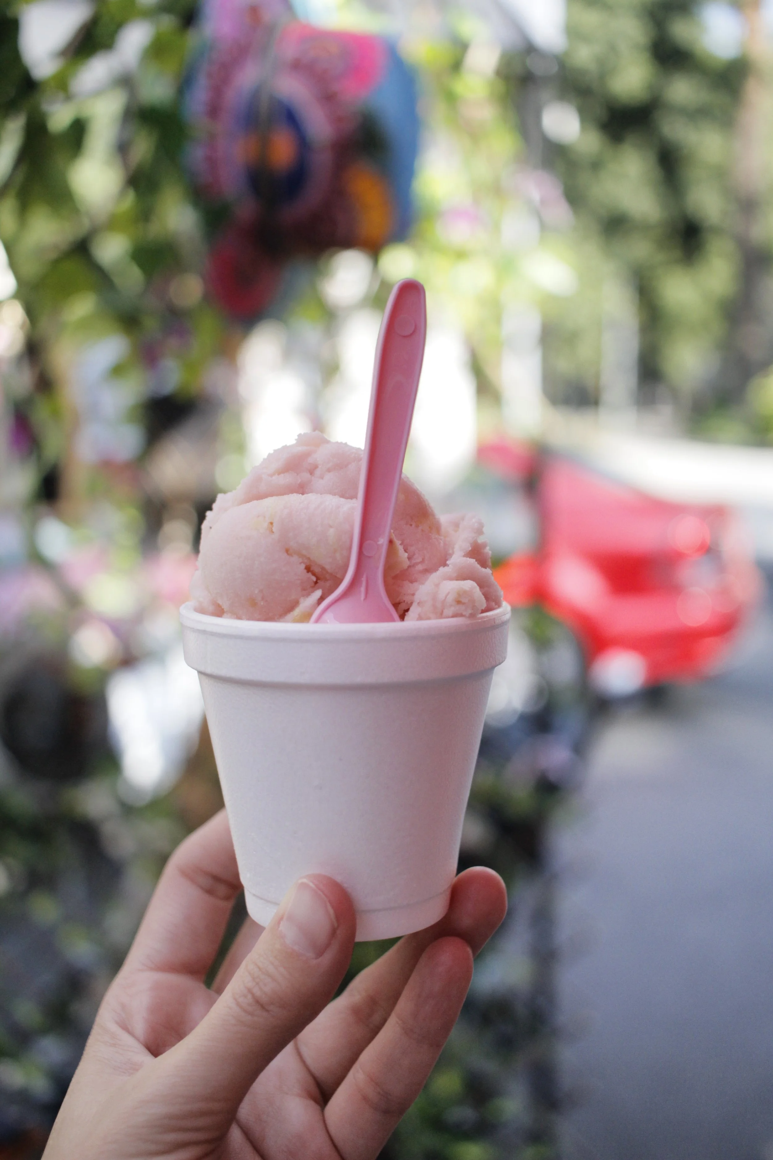 A hand holding a pink spoon in a cup of pink ice cream outdoors with a blurred background of trees and a red vehicle. Mexico City, Mexico.