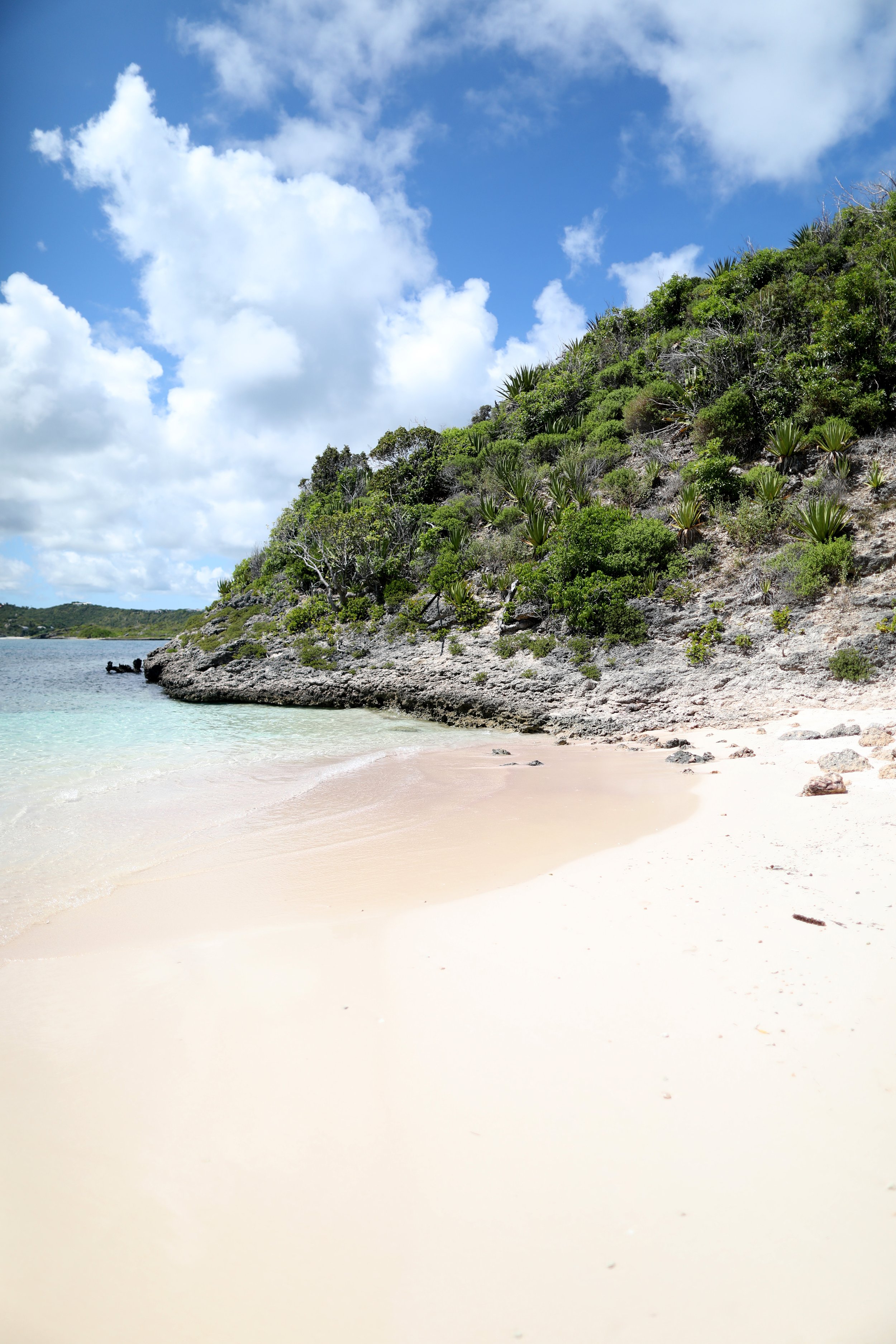 A beach with white sand, a rocky and green hillside, and a partly cloudy blue sky.