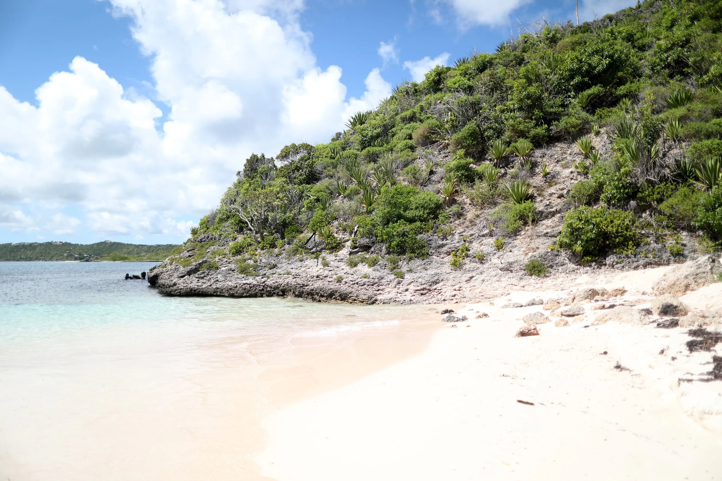 Coastal scene featuring a rocky hill covered with green vegetation, a sandy beach, calm water, and a blue sky with scattered clouds.