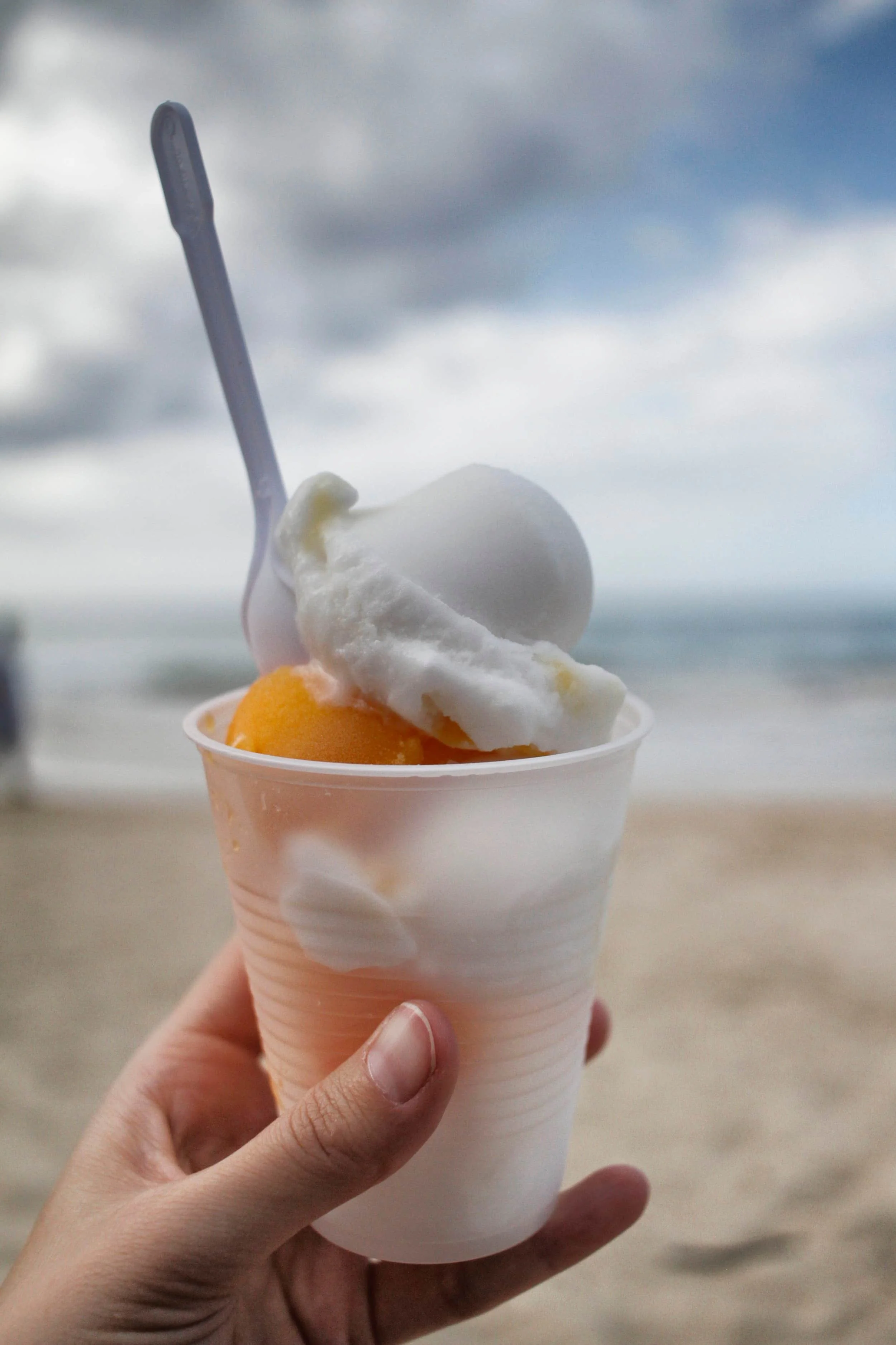 Woman's hand holding a plastic cup with orange ice cream inside with beach in the background in Puerto Rico