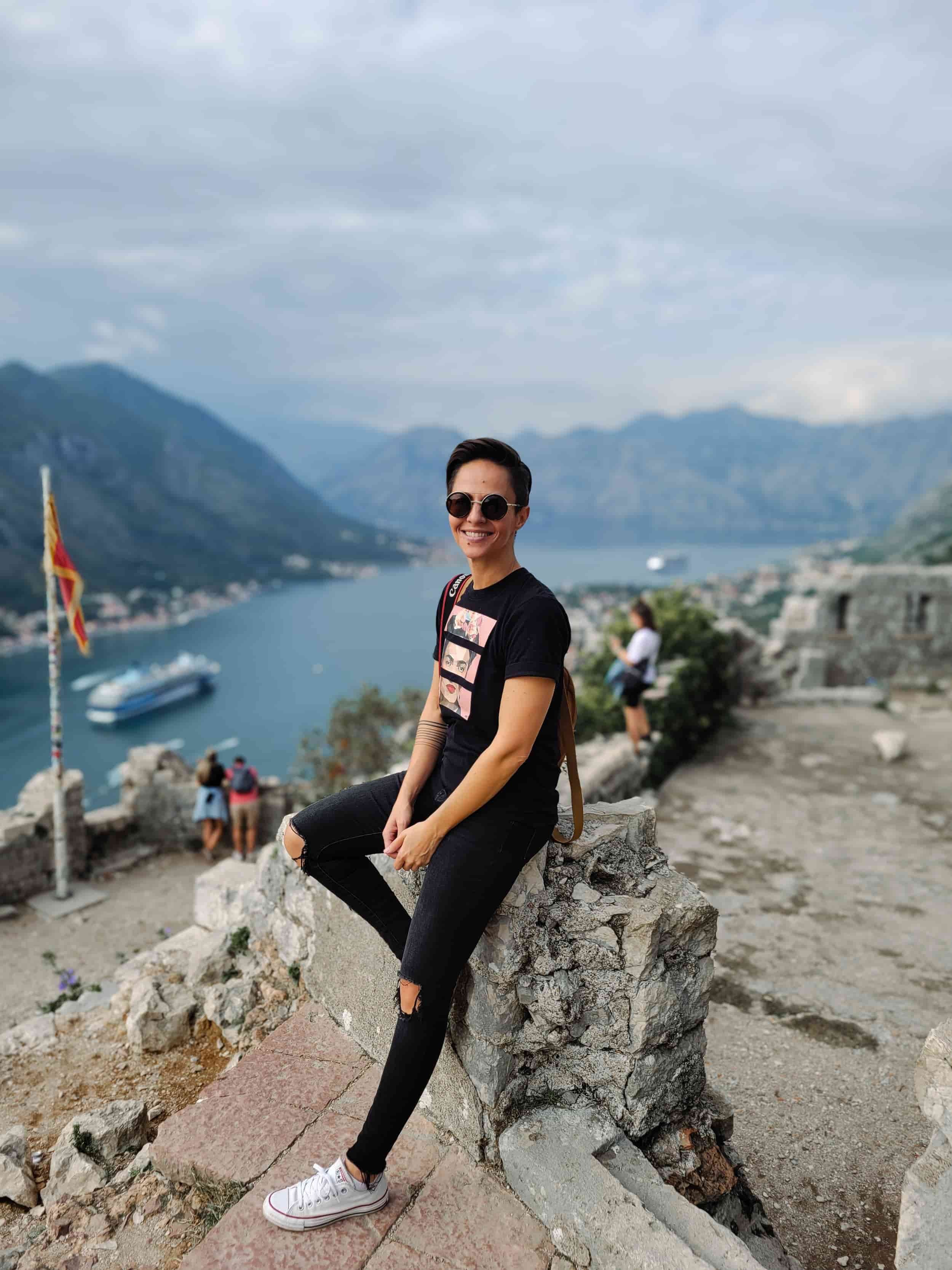 Girl sitting against a rock on a mountaintop with bay and mountains beyond, Kotor, Montenegro