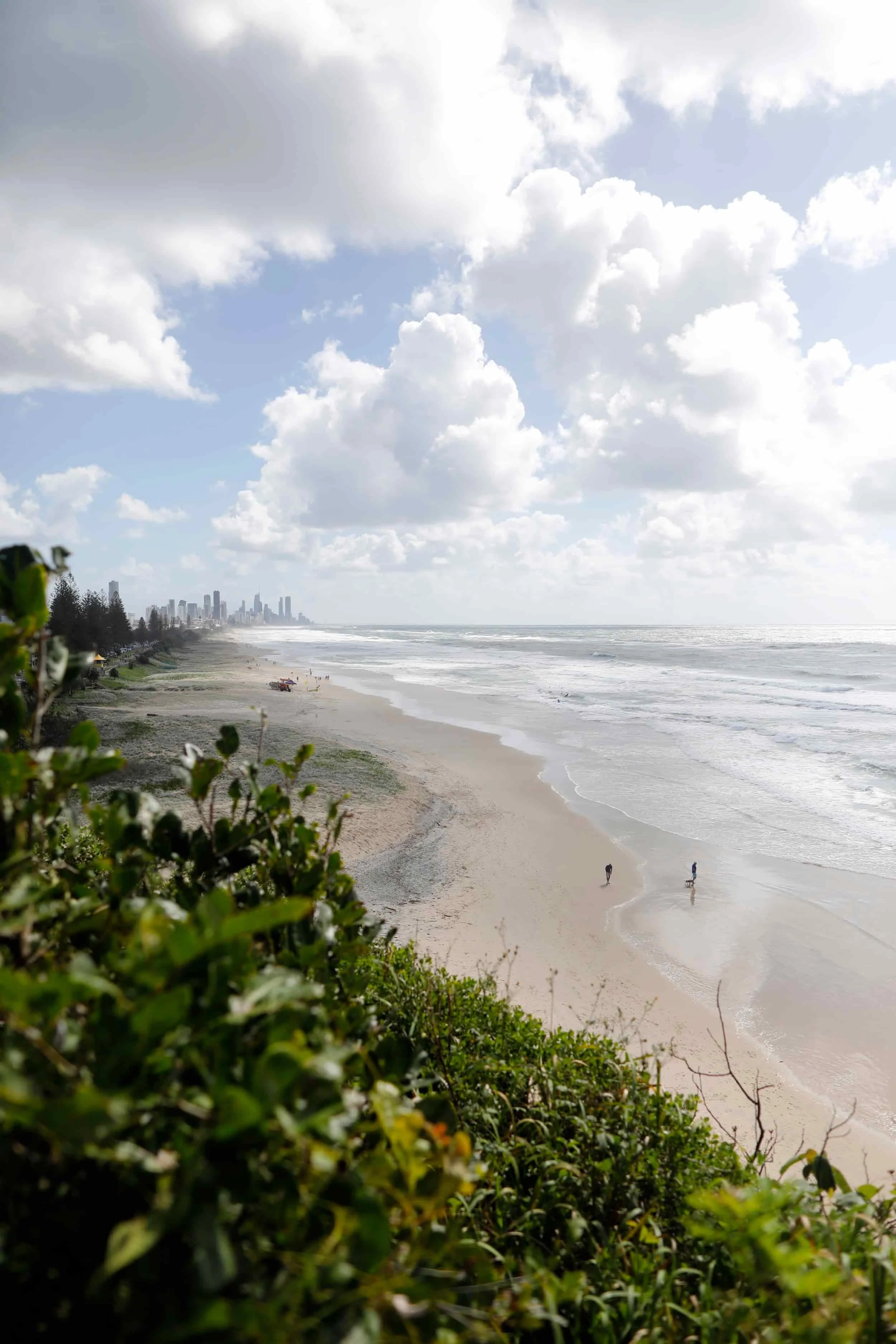 A view of a cloudy sky over a beach with some people walking along the shoreline, with city buildings visible in the distance.