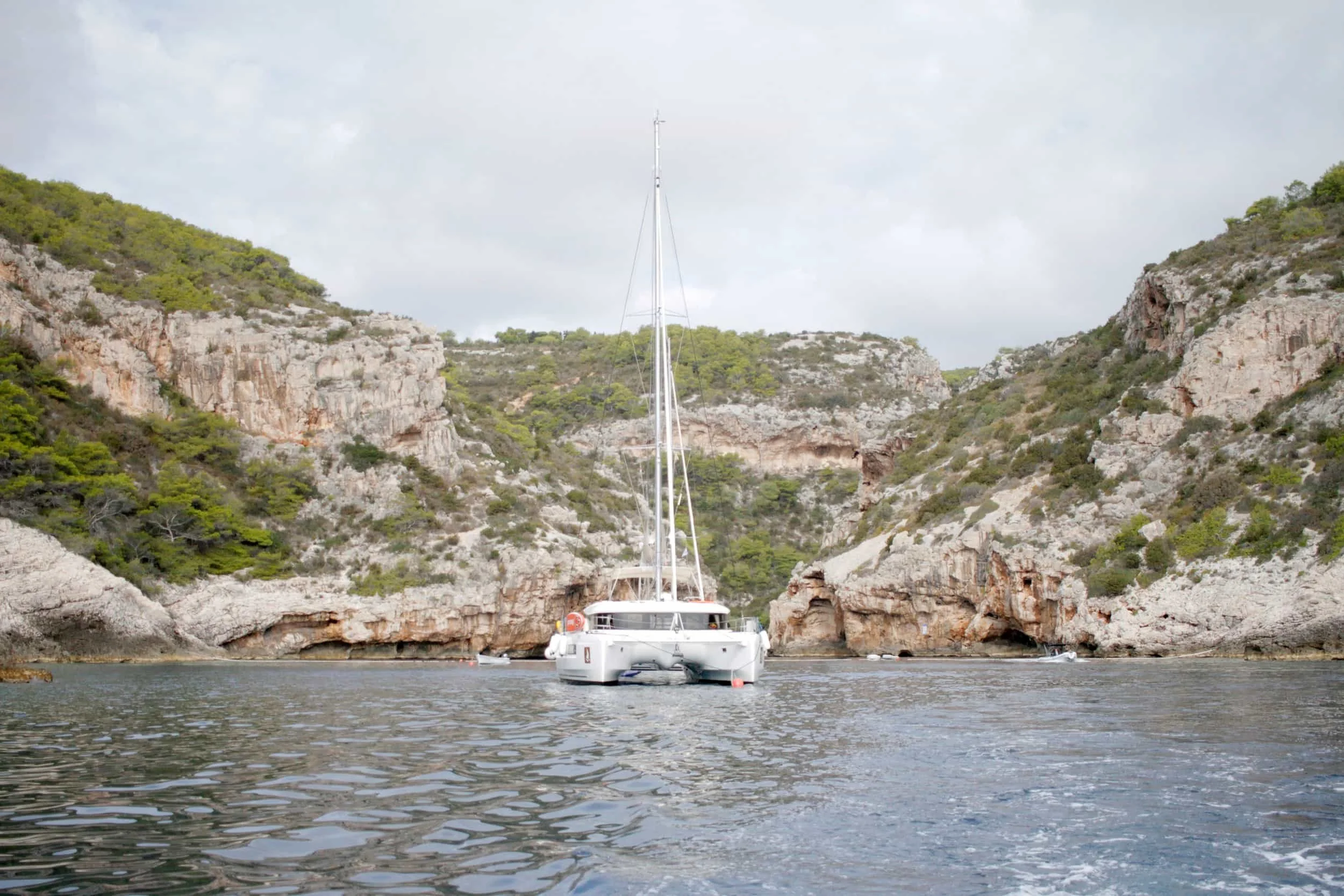 A white sailboat with a tall mast cruising through calm water near rocky cliffs with sparse green vegetation.