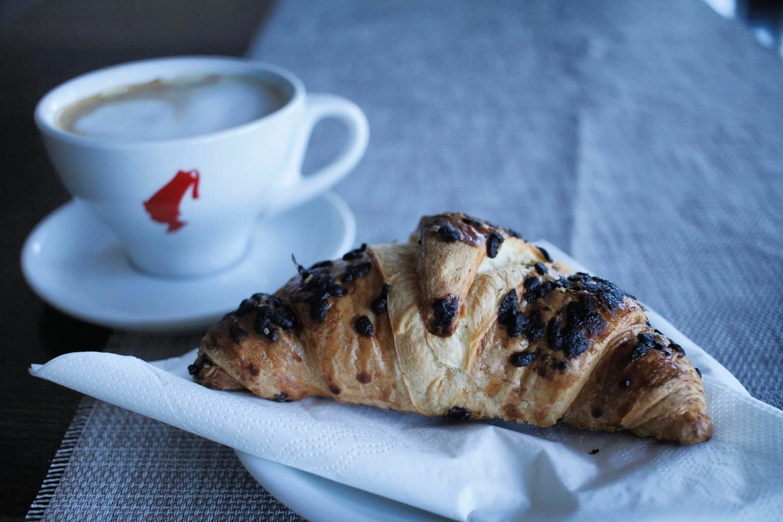 A croissant with chocolate chips on a white paper napkin, alongside a cup of coffee on a saucer with a red logo, on a dark textured table. Bosnia & Herzegovina.