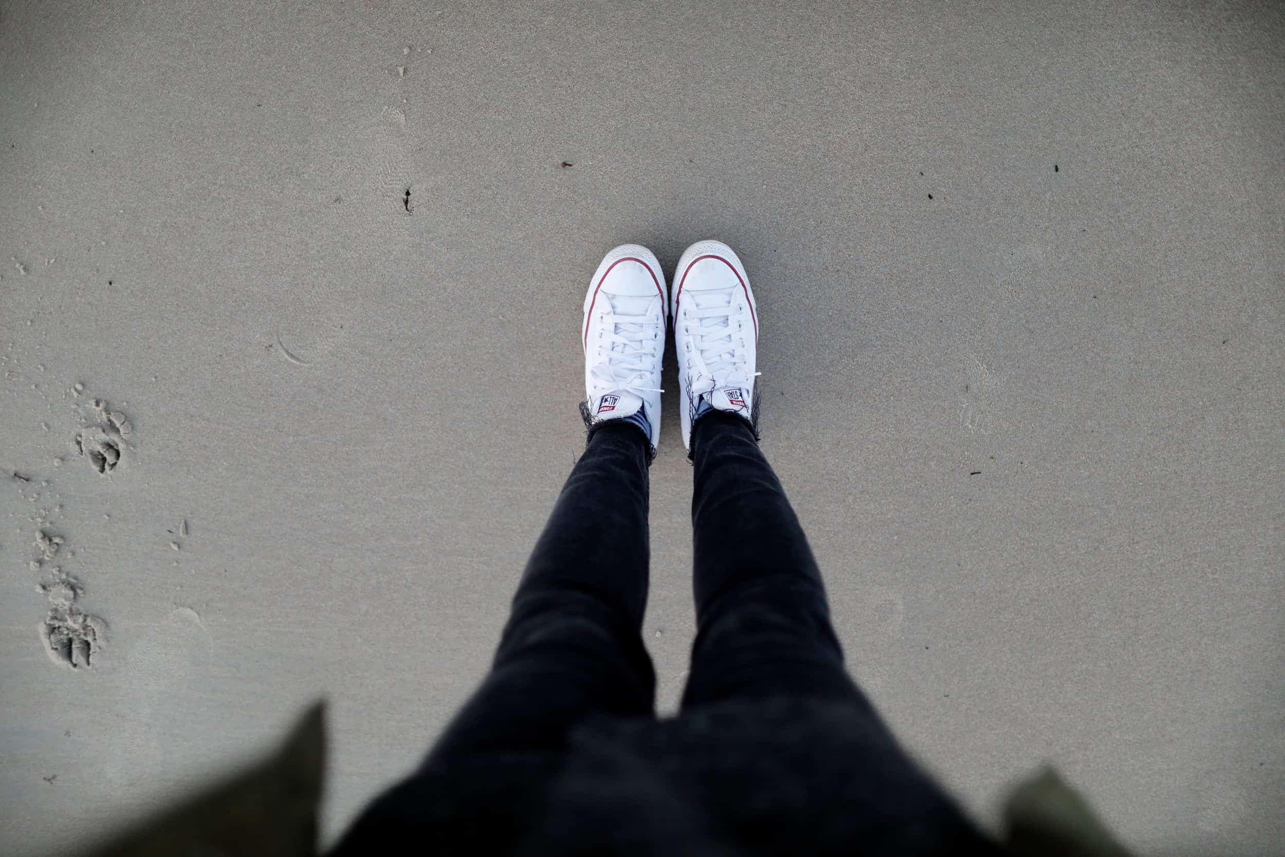 Person wearing white sneakers and black pants standing on sandy beach with footprints.