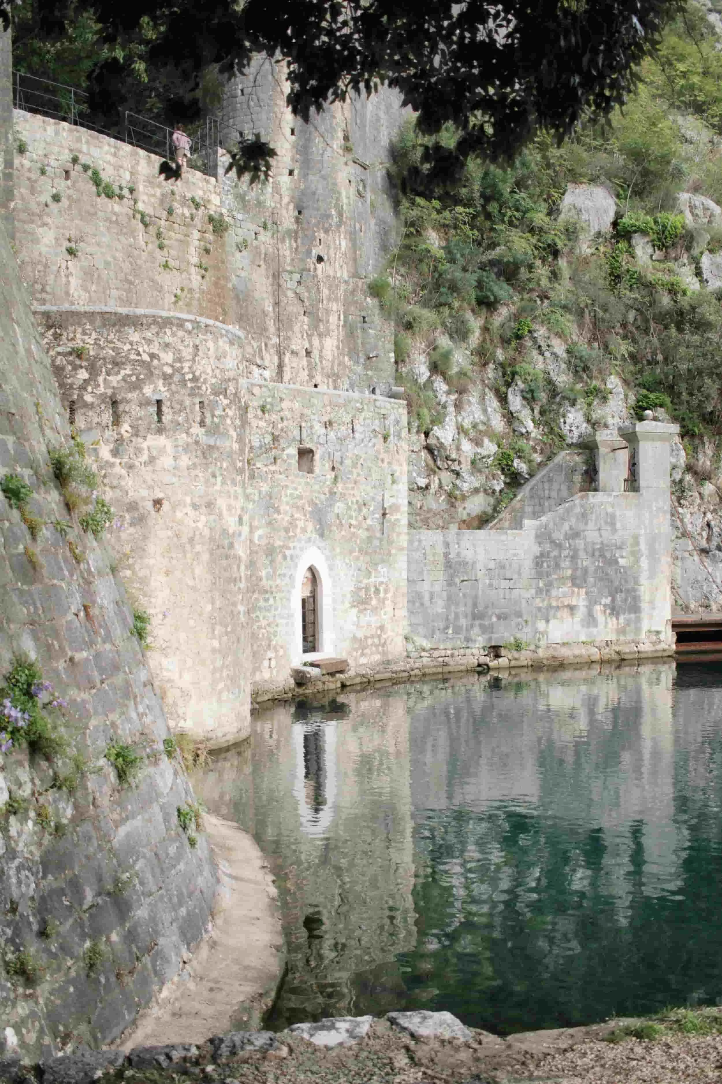 Stone fortress wall with an arched doorway at the water's edge and a pathway along the top of the wall. Trees and rocks surround the area, reflecting in the calm water. Kotor, Montenegro.