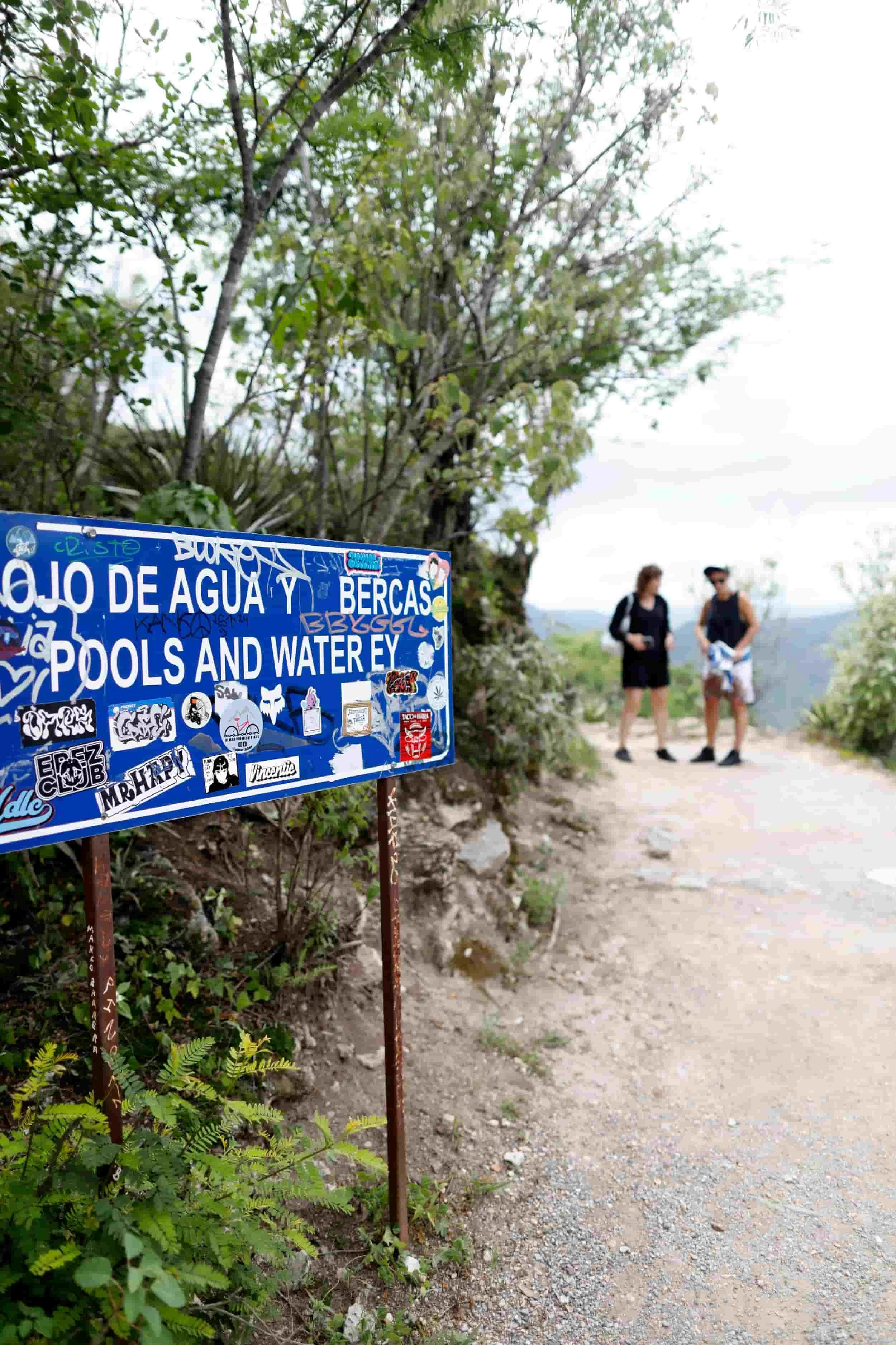Blue sign with graffiti and stickers about water pools, water bodies, and swimming areas, located on a dirt path surrounded by greenery, with two people in casual clothing walking in the background.